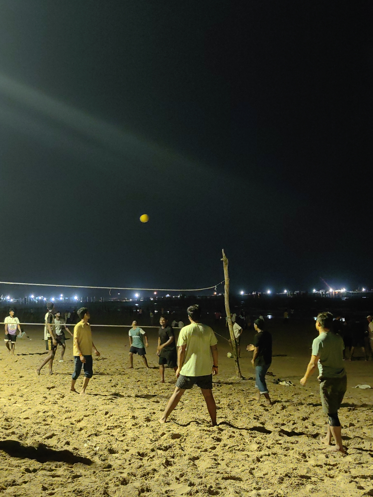 A group of young men are playing volleyball on a sandy beach at night, near the Arabian Sea, likely at Juhu Beach in Mumbai, India. The scene is illuminated by artificial lights, creating strong shadows on the sand. A yellow volleyball is visible in the air above the net. The players are dressed in casual attire, including t-shirts and shorts. The atmosphere appears to be energetic and recreational. In the background, the coastline is dotted with lights, suggesting the presence of a city or town.