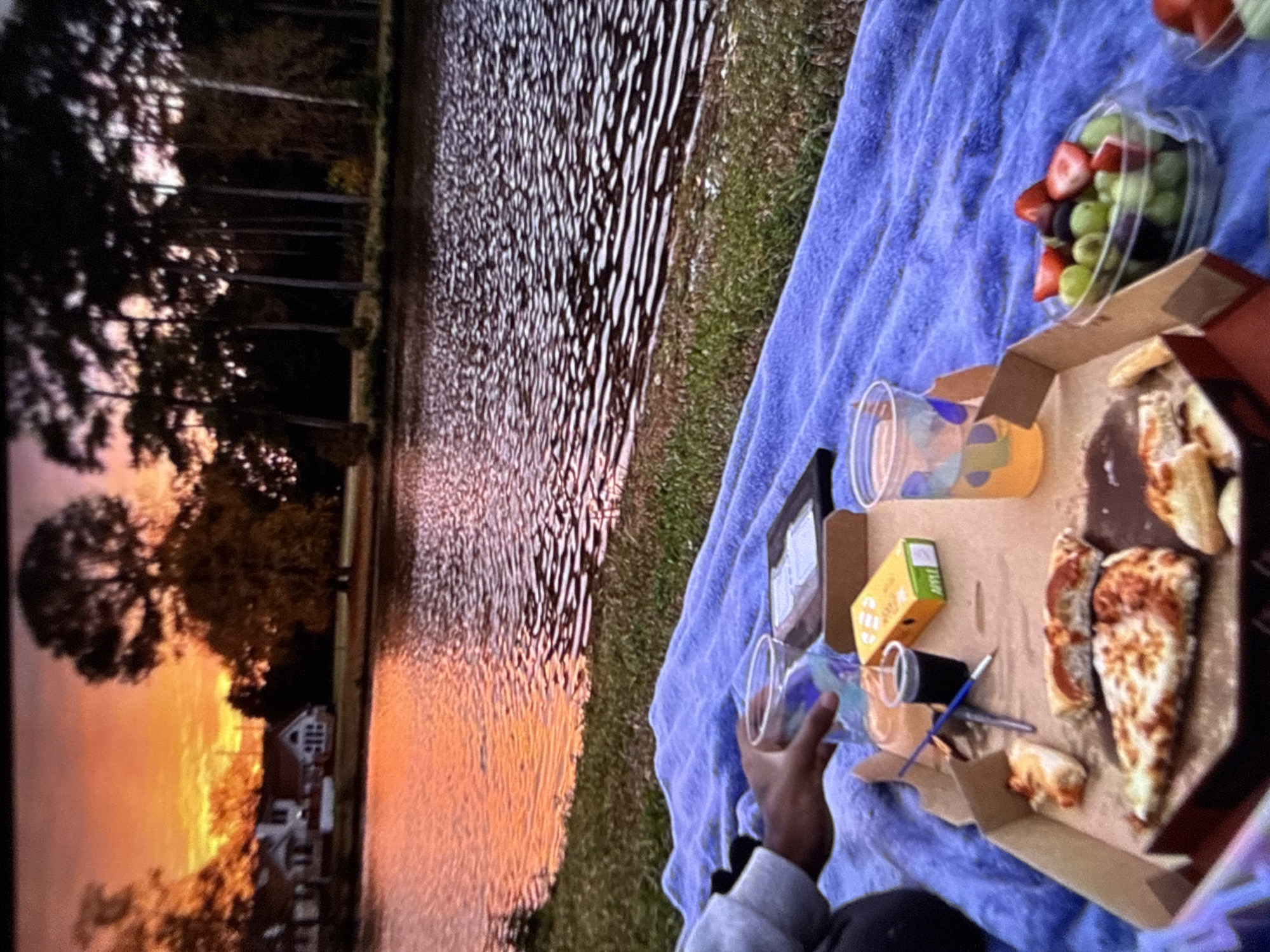 The image depicts a picnic scene by a body of water during sunset. A person's hand is visible, holding a clear plastic cup. In the foreground, a pizza box with several slices of pizza, a small box of what appears to be apple sauce, a small cup of dark liquid, and a paintbrush are arranged on a blue blanket. To the right of the pizza box, a clear plastic container filled with a mix of grapes, strawberries, and possibly blueberries is present, along with another plastic cup containing an orange beverage. The background features a tranquil lake reflecting the warm hues of the sunset, with houses and trees visible on the opposite shore. The overall ambiance suggests a peaceful evening gathering outdoors. The location is not specified to be in Varna, Bulgaria.