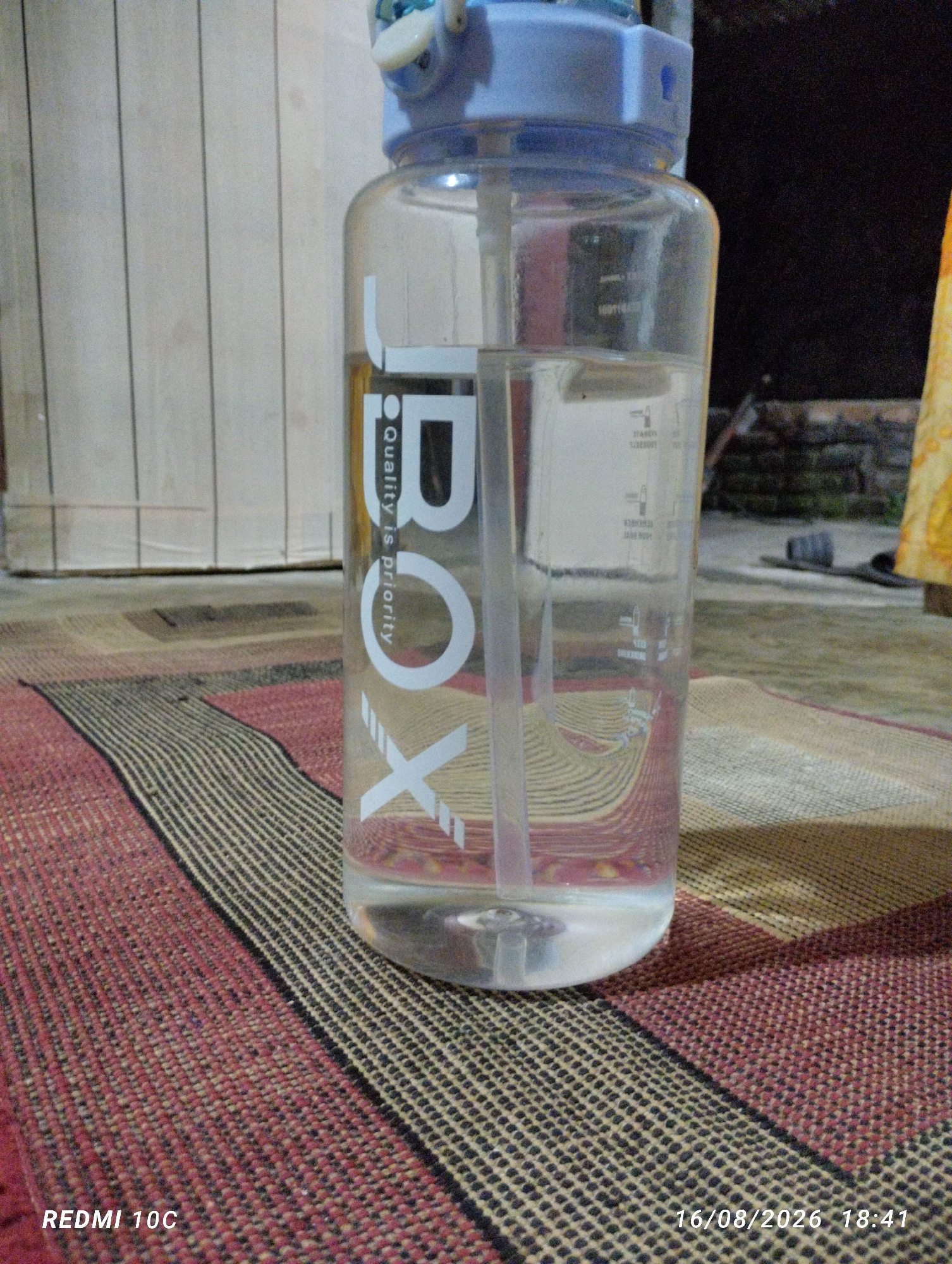 A clear plastic water bottle with a blue lid and a white straw is standing upright on a woven mat with red and black stripes. The bottle has a white logo on it that reads "BOX" vertically, with the text "Quality is priority" in smaller font next to it. The bottle appears to be partially filled with clear liquid. In the background, a wooden wall is visible, and to the right, there is a dark area and a yellow object. The bottom left of the image shows the text "REDMI 10C" and the bottom right shows the date and time "16/08/2026 18:41". The scene is indoors or in a covered outdoor area, as indicated by the wooden wall and the lighting conditions. The location is identified as Kembiritan, Indonesia.