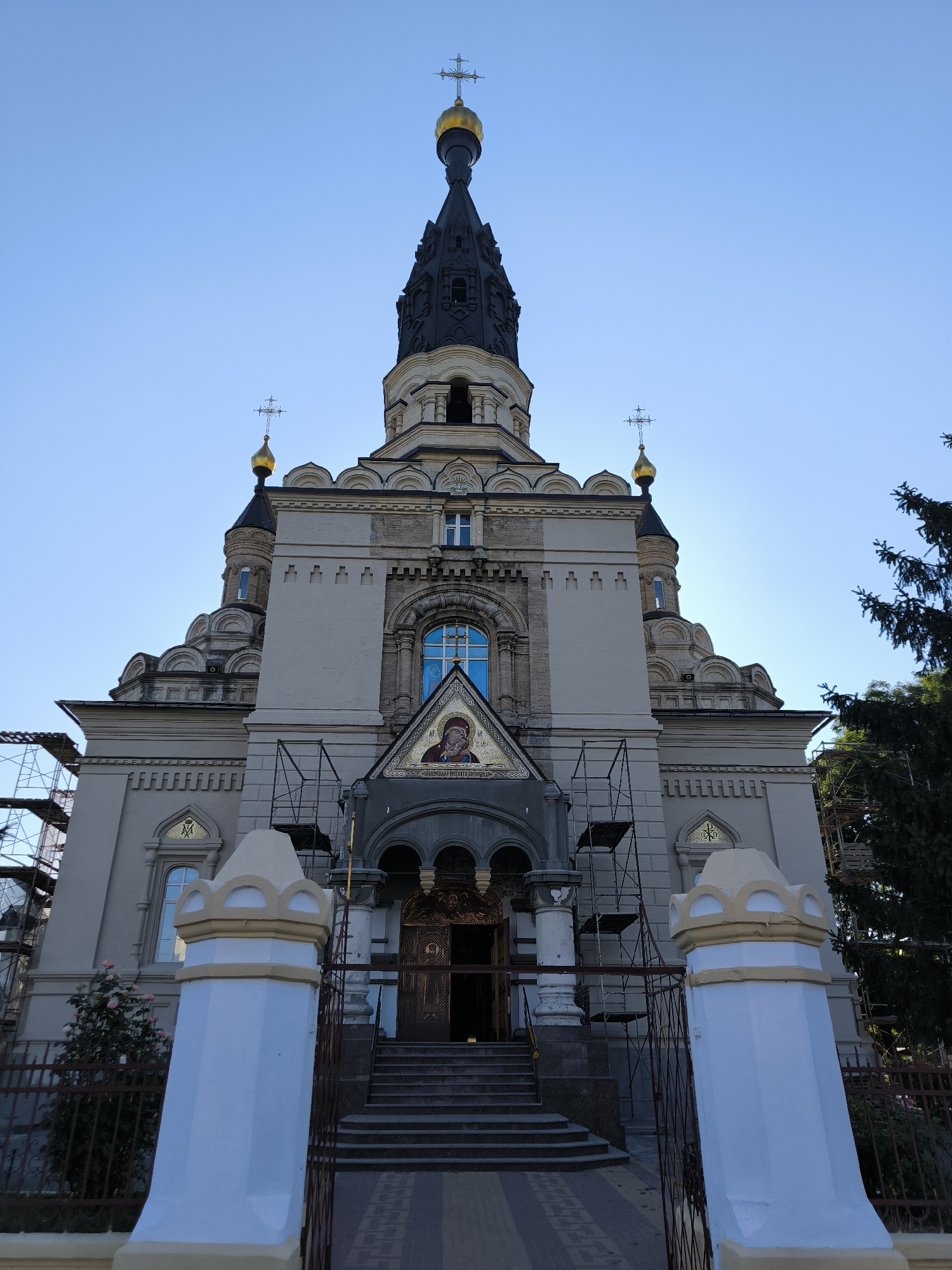 This is a low-angle outdoor shot of a large, ornate Orthodox church, likely under renovation, with scaffolding visible on the left and right sides of the facade. The church is light-colored, with a tall, dark spire topped with a golden dome and cross. There are multiple smaller domes with crosses, and decorative architectural elements such as arched windows and intricate carvings. A mosaic icon of a religious figure is prominently displayed above the entrance. In the foreground, two light-colored, decorative pillars stand behind a metal fence. A tree with dark foliage is visible on the right side of the frame. The sky is clear and blue. The church is St. Nicholas Cathedral in Mykolaiv, Ukraine.