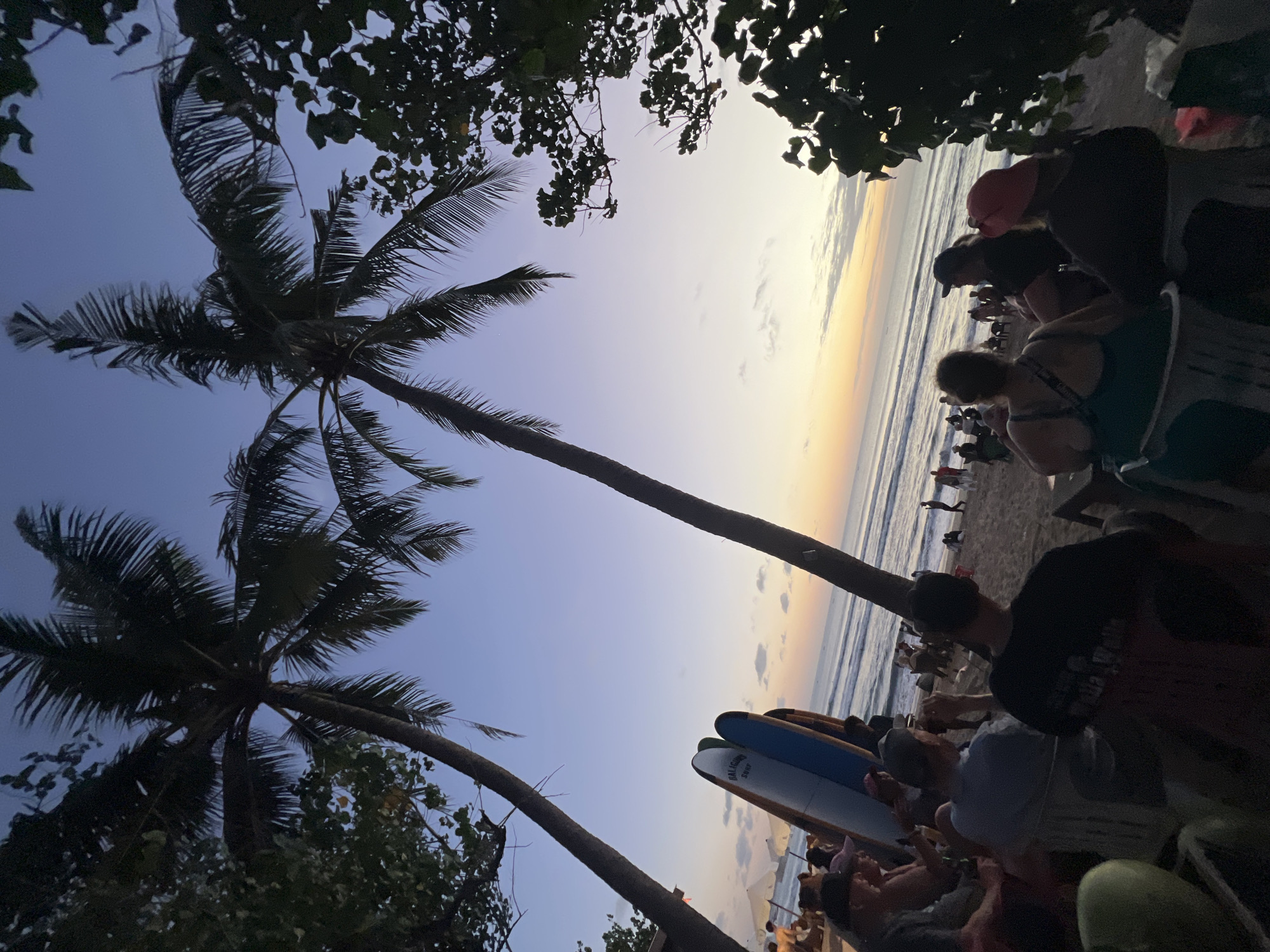 The image depicts a beach scene in Kuta, Indonesia, during twilight. Two prominent palm trees frame the view of the ocean and the sky, which is transitioning from a deep blue to hues of orange and yellow near the horizon. Several surfboards are propped up on the sandy beach, suggesting a location popular for surfing.
In the foreground, a group of people are seated in chairs on the sand, facing the ocean. They appear to be socializing and watching the sunset. Their clothing is casual, fitting for a beach setting. Some individuals are wearing hats, and one person is holding a drink. The overall atmosphere seems relaxed and convivial.
Further down the beach, more people are visible, some standing and walking along the shoreline, while others are seated or gathered in small groups. The waves are gently rolling onto the beach. The presence of umbrellas and chairs indicates a developed beach area, likely with amenities for visitors. The surrounding foliage, including the leaves of the palm trees and other trees on the edge of the scene, adds to the tropical ambiance.