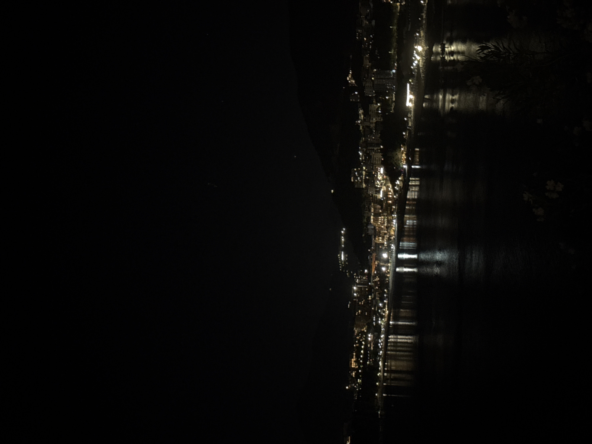 The image depicts a nighttime scene of the city of Rafailovići, Montenegro, as viewed from an elevated position. The city is situated along a body of water, likely the Adriatic Sea, with city lights reflecting on the water's surface. The left side of the image is dominated by darkness, suggesting the presence of a hillside or mountain. On the right side, city lights are visible along the coast. No people are identifiable. The environment is dark and still.
