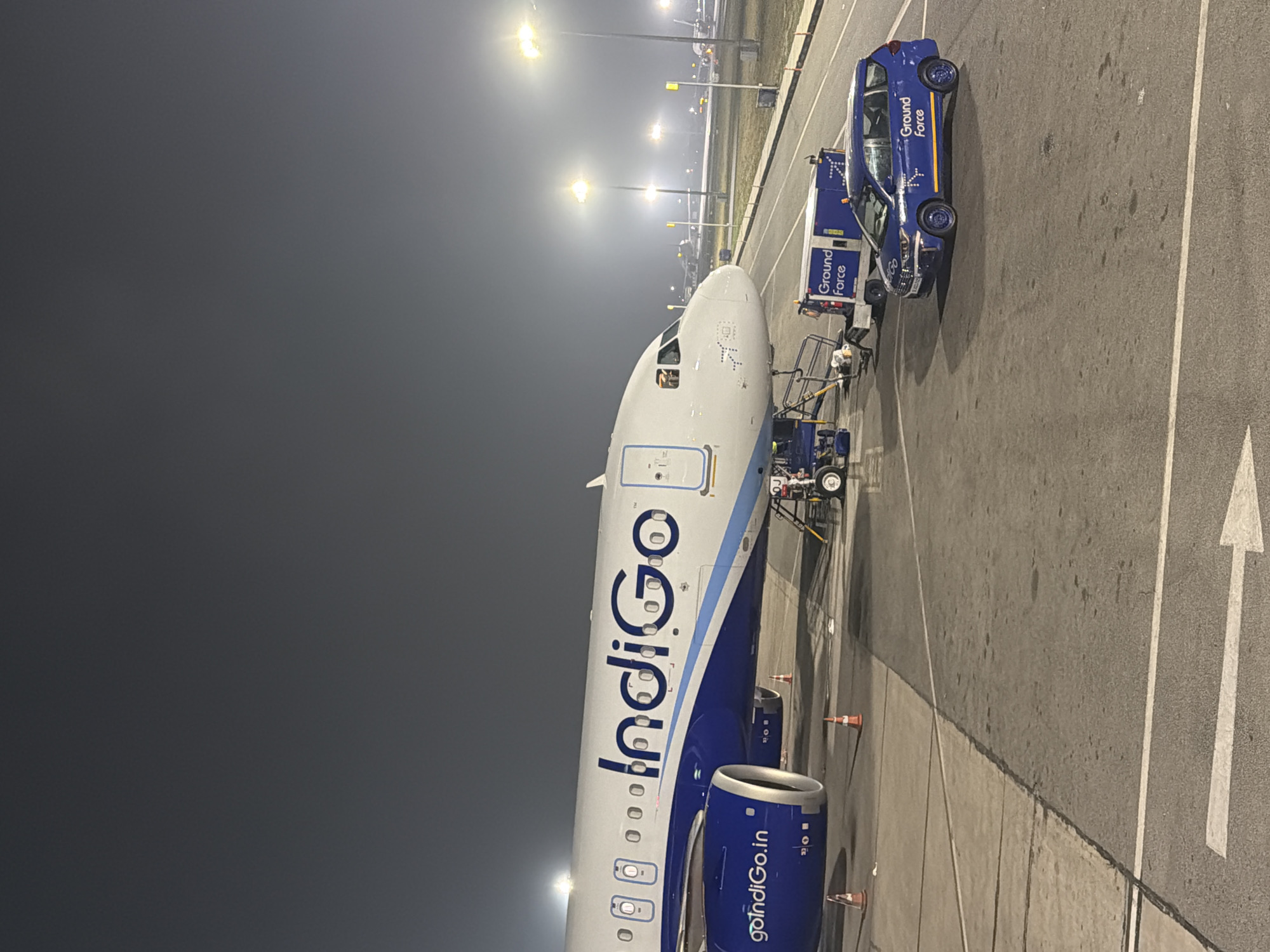 An Indigo airline airplane is parked on the tarmac at Indira Gandhi International Airport in New Delhi, India. The airplane is white with blue accents and the Indigo logo prominently displayed on its side. A blue ground vehicle with "Ground Force" written on it is parked next to the airplane. The scene appears to be at night, with airport lights illuminating the area. The sky is dark and overcast. The ground is paved with asphalt and concrete, with markings indicating taxiways and parking areas. There are several orange traffic cones placed around the airplane. The overall environment suggests an active airport setting at night.