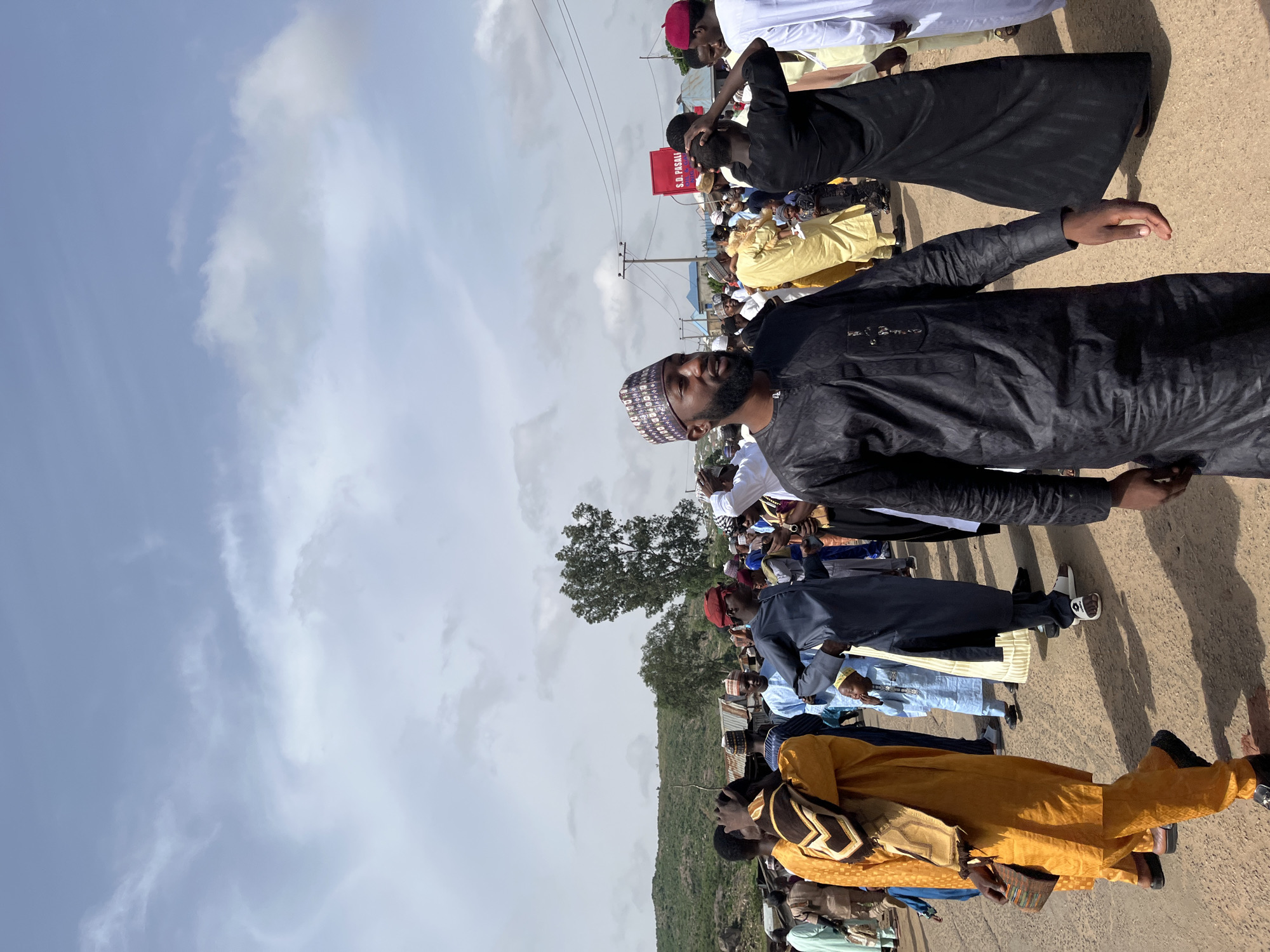 A crowd of people, predominantly men, stands outdoors on a dirt or paved surface under a sky with light clouds. The location is suggested to be Jos, Nigeria.
The individuals are dressed in a variety of clothing, ranging from traditional attire with varying colors like yellow, blue, black, and white, to more modern casual wear. Some wear caps or hats.
Behind the crowd, vegetation is visible, suggesting the presence of trees and possibly a hillside. Utility poles are also visible, indicating a built environment. A sign with the text "S.D. PASALI" is positioned near the crowd.