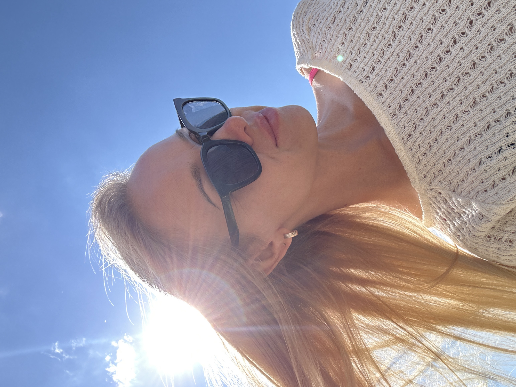 A woman with blonde hair, wearing sunglasses and a cream-colored sweater, is pictured outdoors against a bright blue sky. The sun is shining, creating lens flare and highlighting her hair. She is looking away from the camera, to the right. The scene is not identifiable as being in the city of Приморск, Russia.