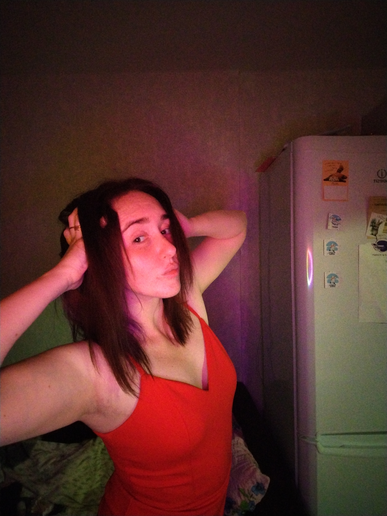 A woman with dark brown hair is standing indoors, wearing a red tank top. She has her hands behind her head and is looking directly at the camera with a slight pout. Behind her is a beige wall illuminated with pink lighting. To the right of the frame is a white refrigerator covered with magnets. The location is possibly a residential setting within the city of Izhevsk, Russia.
