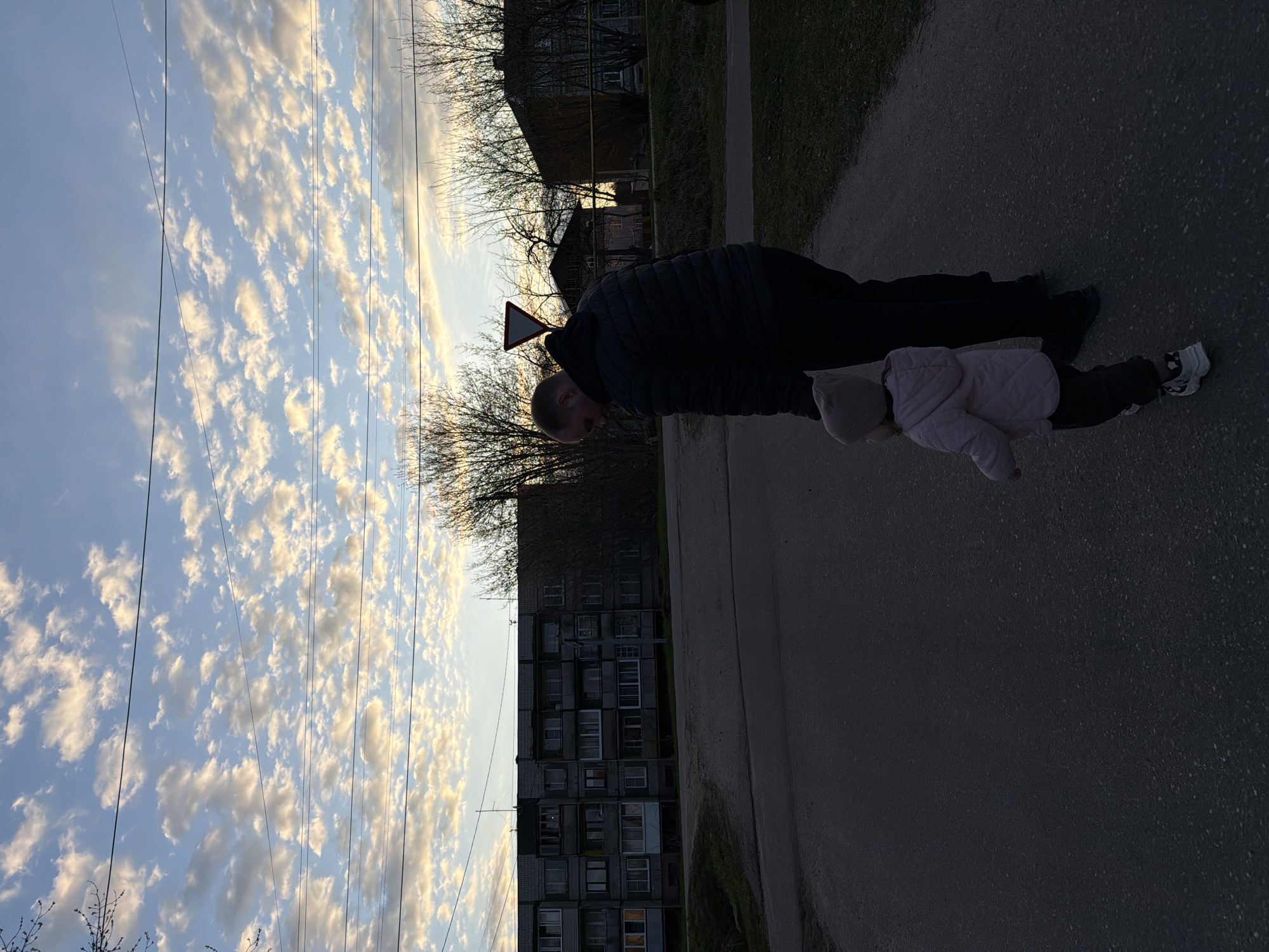 The image depicts a man and a small child walking on a paved street. The man is wearing a dark jacket. The child, wearing a light pink jacket and hat, is holding the man's hand. They are walking next to a grassy area and a large, block-style apartment building. Overhead, power lines stretch across a sky filled with numerous small, puffy clouds. A triangular road sign is visible in the background. The location is likely Reshetnikovo, Russia, judging by the appearance of the apartment buildings and the general landscape.