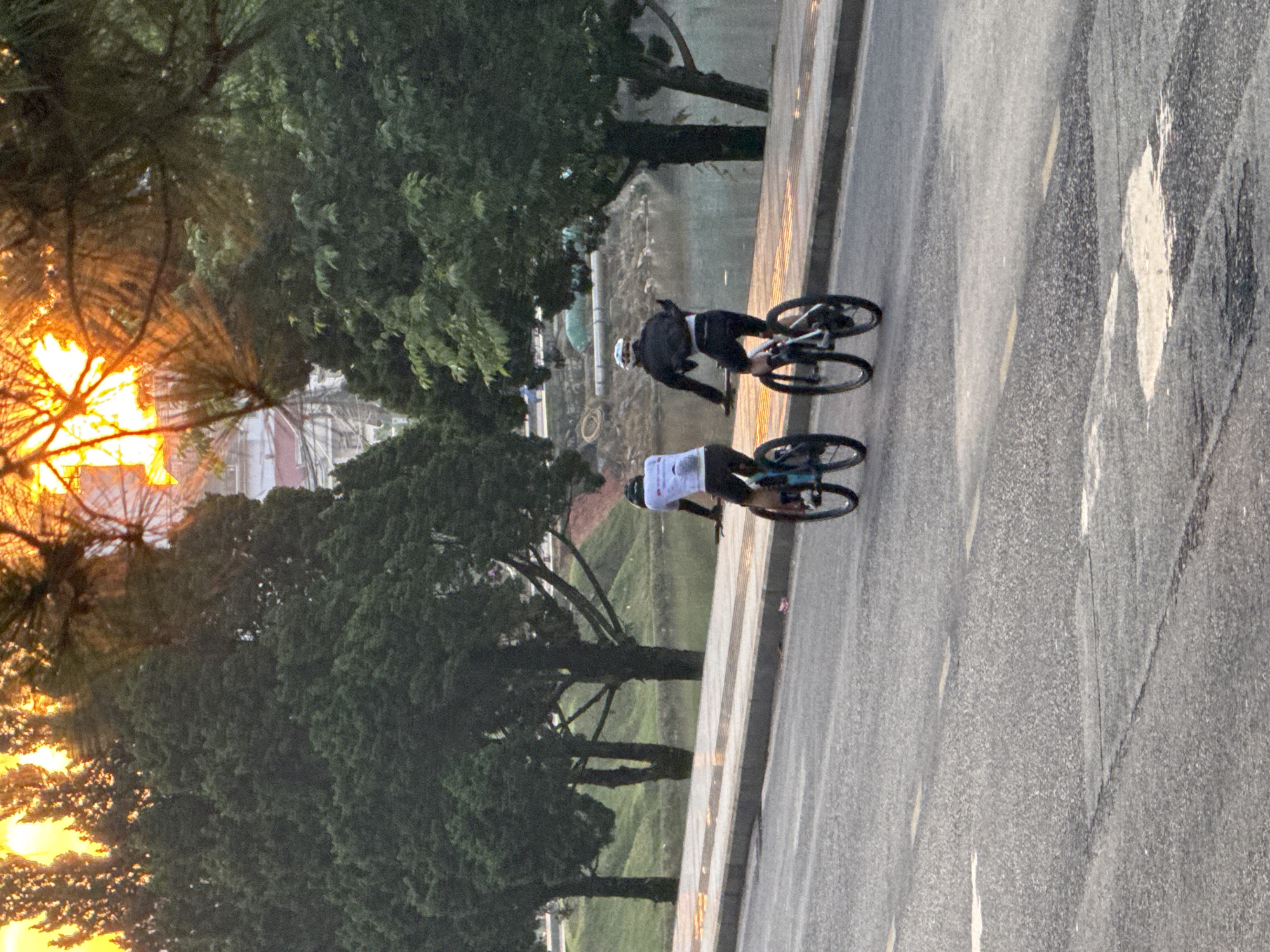 Two individuals are cycling on a paved road in Lâm Viên - Đà Lạt, Vietnam, during what appears to be sunrise or sunset, as indicated by the warm, bright light filtering through trees in the background. The road has yellow lane markings and some patches of water or wetness. To the left of the road is a sidewalk with a curb, and beyond that, a body of water, possibly a lake or pond, with grassy slopes and trees. Further in the background, more trees and some buildings are visible, with the sun's rays creating a dramatic effect through the foliage. The cyclists are both wearing helmets and athletic clothing, with one in a white shirt and black shorts, and the other in a black jacket and shorts. They are moving away from the camera, suggesting a forward momentum in their cycling journey. The overall atmosphere is peaceful and natural, with a hint of urban development in the distance.
