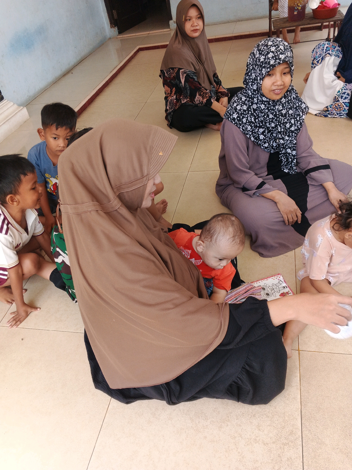 In Kadu, Indonesia, a group of children and women are gathered on a tiled floor, appearing to be in an indoor setting with a light blue wall and doorway visible in the background. A woman in a brown hijab is in the foreground, seated and facing a young child in an orange shirt. The child is looking down at a book. To the left of the woman in brown, a young boy in a blue shirt is looking towards the camera. To the right of the woman in brown, another child in a pink shirt is also seated on the floor, partially visible. In the background, a woman in a black and white patterned hijab is seated, and another woman in a light purple outfit and a white hijab is also visible. The scene suggests a communal gathering, possibly for a lesson or storytelling session.