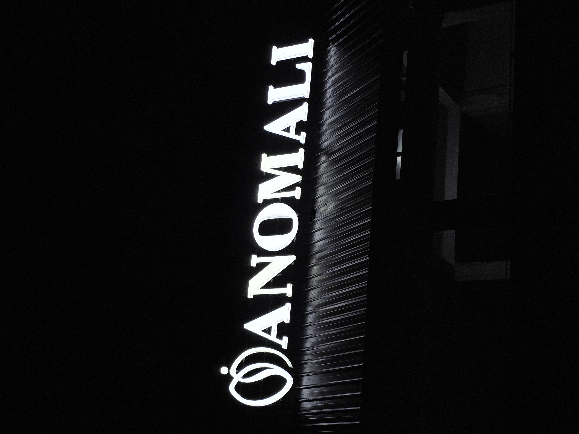 A black and white, low-angle, eye-level shot captures a white illuminated sign against a dark background. The sign, mounted on a corrugated metal roof, spells out "ANOMALI" in capital letters. To the left of the text is a stylized logo resembling two intertwined rings with a small dot above, potentially representing a human figure. The lighting of the sign creates a stark contrast with the dark, indistinct surroundings, suggesting it is nighttime or in a dimly lit area. The corrugated roof has vertical lines, indicating a sloped surface. The lower portion of the image is predominantly black, with faint outlines of what appears to be a structure beneath the roof. The scene is located in Uleeue, Indonesia.