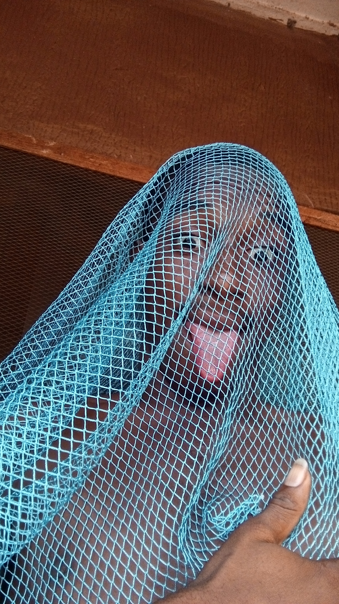 A person with dark skin is partially visible behind a light blue mesh netting. The person is sticking their tongue out. A brown wooden structure is visible in the background. The location is unspecified, and no information indicates the city of Onitsha, Nigeria. A hand with dark skin is also partially visible, holding the netting.