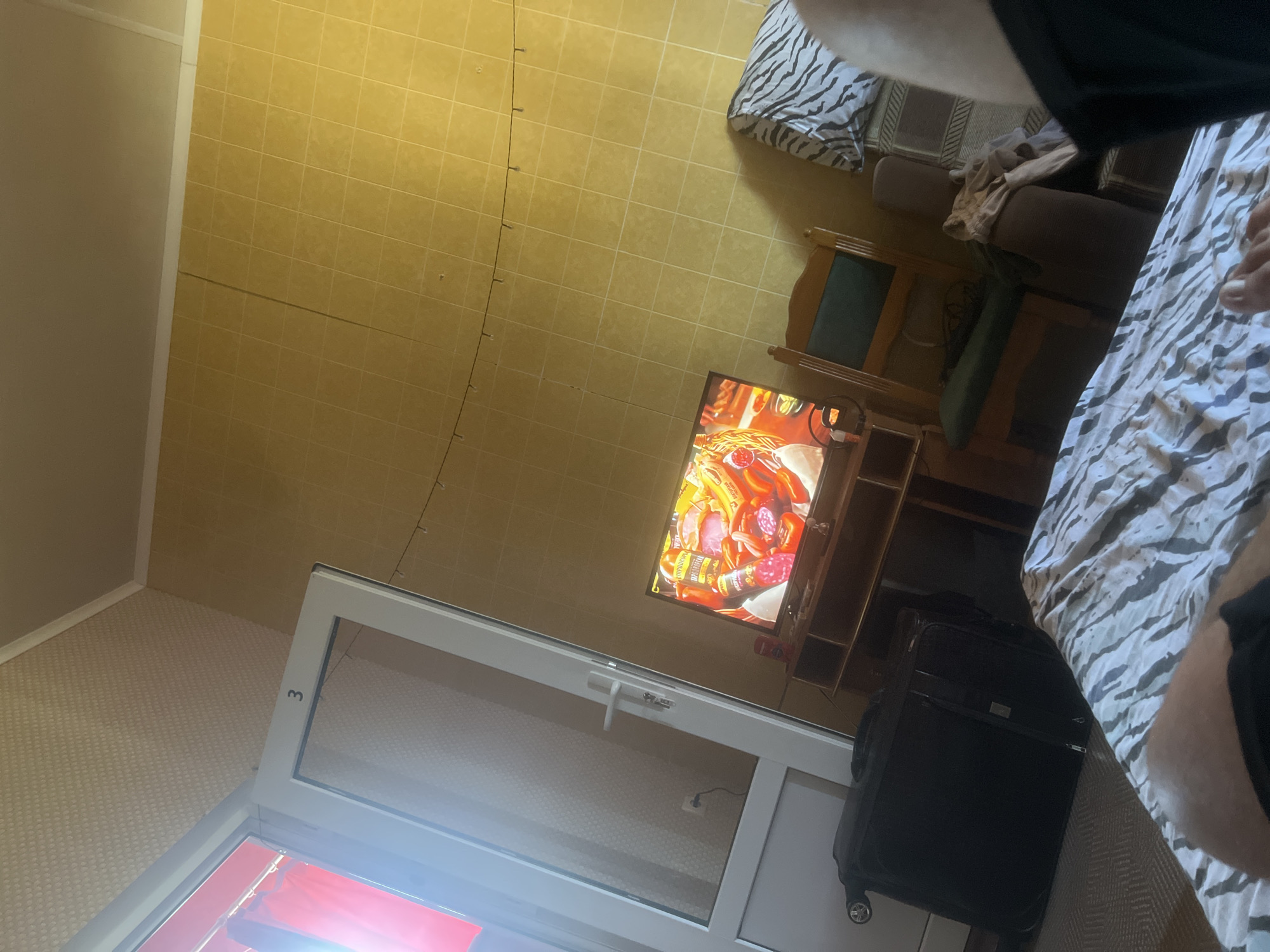 The image shows a room with yellow tiled walls. In the foreground, a bed with a black and white striped duvet is visible. A person's legs are on the bed. To the left, a black suitcase with wheels stands on the floor. A white door with the number "3" on it is ajar, revealing a red curtain. In the middle ground, a television is on, displaying an image of various types of sausages and peppers. A wooden TV stand is below the television. A wooden chair with a green seat is next to the TV stand. To the right, a part of a sofa with a zebra-patterned pillow is visible. A string of small lights is hung across the upper portion of the wall. The overall impression is that of a simple hotel room or guest house.