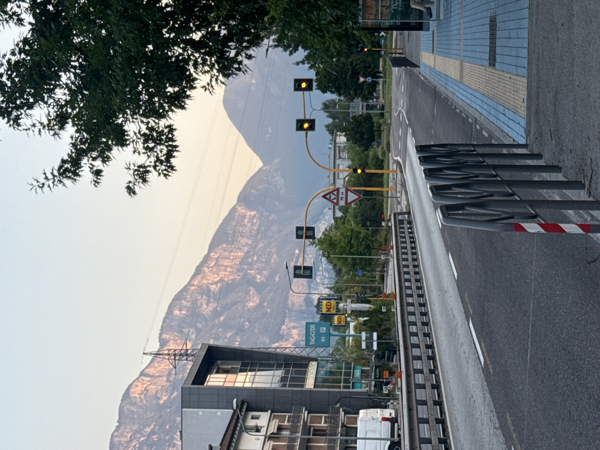 The image depicts a street scene in Trento, Italy, with a focus on a road leading towards a roundabout. In the foreground, a grey metal barrier with red and white striped markings lines the side of the road. The road itself is asphalt with white lane markings. To the right of the road, a pedestrian walkway is visible, paved with blue and yellow tiles. Further in the background, traffic lights with yellow signals are illuminated, indicating caution. Buildings, including a modern structure with glass windows and older residential buildings, are situated on the left side of the street. Mountainous terrain, illuminated by the soft light of dawn or dusk, forms the backdrop. Trees are present on the right side of the image, partially obscuring the view of the sky. A white van is parked on the left. The overall atmosphere suggests a quiet urban environment on the outskirts of the city, with the natural landscape of the mountains serving as a prominent feature.