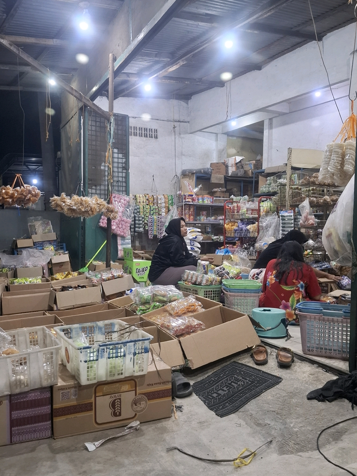 The image depicts a dimly lit interior of what appears to be a small grocery store or market stall in the city of Medan, Indonesia. Several people are present, some of whom are engaged in activities related to the shop. In the foreground, cardboard boxes are stacked, some open and containing various packaged goods. Several plastic baskets, filled with what seems to be snacks or other merchandise, are also visible.
Two women are seated on the floor among the merchandise. One woman, wearing a dark hijab and black jacket, is facing left, seemingly observing something. Another woman, wearing a red shirt with a floral pattern, is seated on the floor facing away from the camera, reaching into a cardboard box. A third person, also wearing a dark hijab, is partially visible behind the first woman.
The environment is cluttered, with shelves filled with more products visible in the background. Bags of snacks are hanging from the ceiling. The floor is concrete and appears somewhat worn. Lighting comes from overhead bulbs, casting strong illumination and shadows, suggesting it may be evening or the interior is not well-lit by natural light. The overall impression is of a busy, informal retail setting.