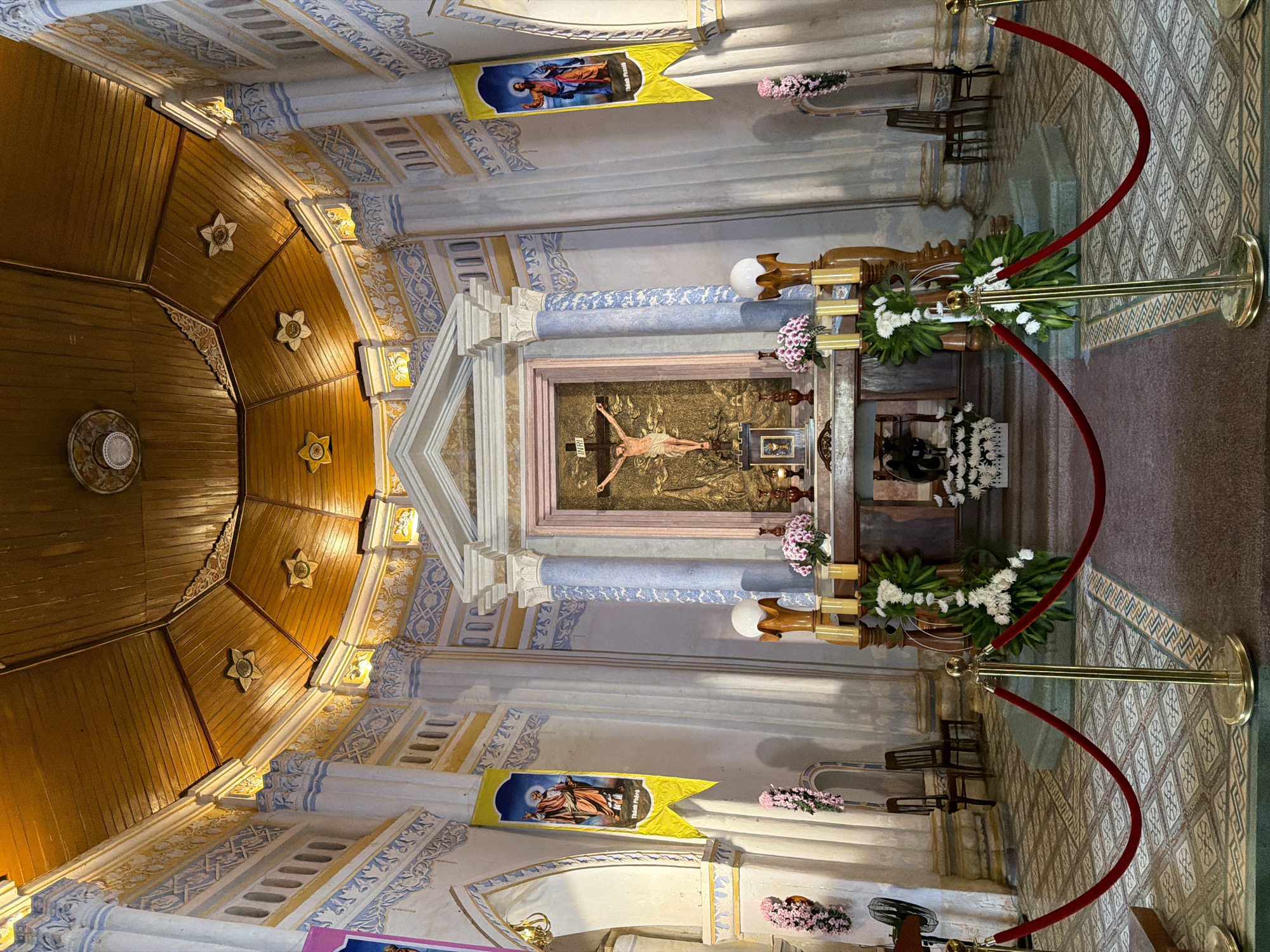 The image depicts the interior of a church in Hội Tín, Vietnam, focusing on the altar area. The altar itself is ornate, with a large crucifix of Jesus Christ prominently displayed at its center, set against a gold backdrop. To the sides of the altar are decorative floral arrangements and religious banners featuring figures of saints. The church's architecture is characterized by white walls with intricate blue and yellow decorative patterns, supporting a wooden, vaulted ceiling. Pillars with Corinthian-style capitals are present, and the floor is tiled with a geometric pattern. Red velvet ropes on golden stanchions delineate a path in front of the altar, suggesting a restricted area for visitors. The lighting appears warm, coming from the ceiling and possibly from unseen sources, highlighting the rich textures and colors of the space. No people are visible in the scene.