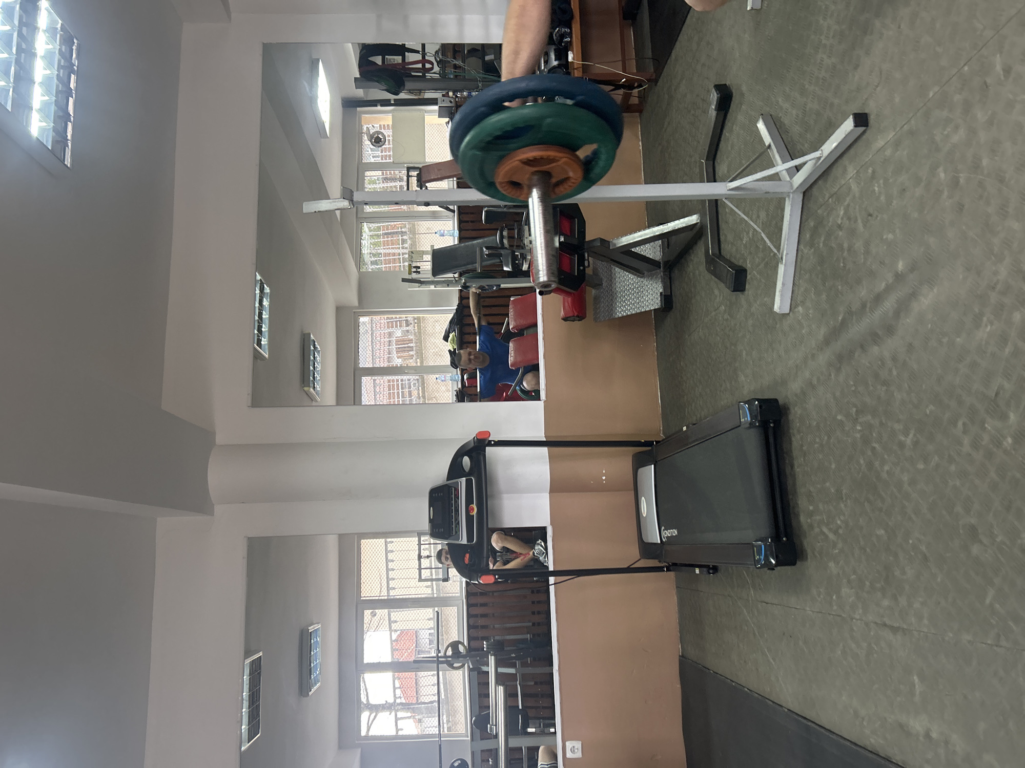 Location: Zlatograd, Bulgaria.
Scene: Interior of a gym.
Objects: Treadmill, weight rack with barbell loaded with weight plates, weight bench, mirrored wall.
People: A person is visible on the treadmill, and another individual is seated on the weight bench in the mirrored reflection.
Environment: The gym is well-lit, and the floor is covered in a grey, textured material. Windows are visible in the background.