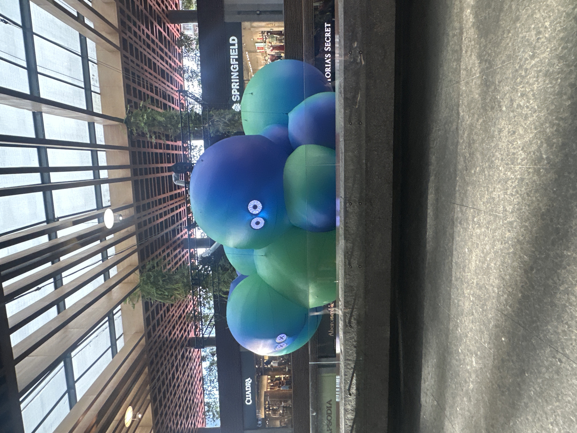 The image depicts a large, inflatable, multi-segmented sculpture resembling a creature with large, cartoonish eyes. The sculpture is predominantly blue and green, with varying shades and gradients. It is situated outdoors, in what appears to be a public space or shopping area, as indicated by the storefronts visible in the background, including "QUADRA," "SPRINGFIELD," and "VICTORIA'S SECRET." Above the sculpture and storefronts, a modern architectural canopy with wooden beams and translucent panels is visible. The sculpture is placed in front of a grey stone ledge. The ground in the foreground is a textured grey surface. The lighting suggests it is daytime. This scene is located in Puebla, Mexico, likely within a commercial or entertainment district. No people are clearly visible interacting with the sculpture, though some figures can be seen inside the storefronts.