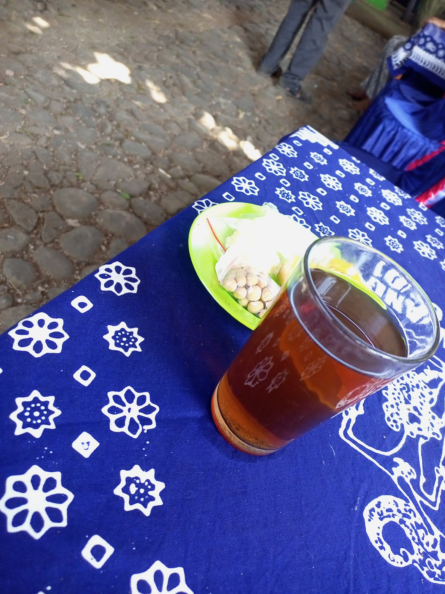 An outdoor cafe table is set with a glass of dark liquid, likely tea, and a small plate of snacks. The table is covered with a vibrant blue cloth featuring a white floral pattern. In the background, people are visible, one person in dark pants and another in a blue skirt with a decorative white top, suggesting a gathering or event. The ground is paved with cobblestones, and dappled sunlight suggests it's daytime. The location is the outdoor seating area of a cafe or eatery in Jatisrono, Indonesia, where patrons can enjoy refreshments in a relaxed atmosphere.