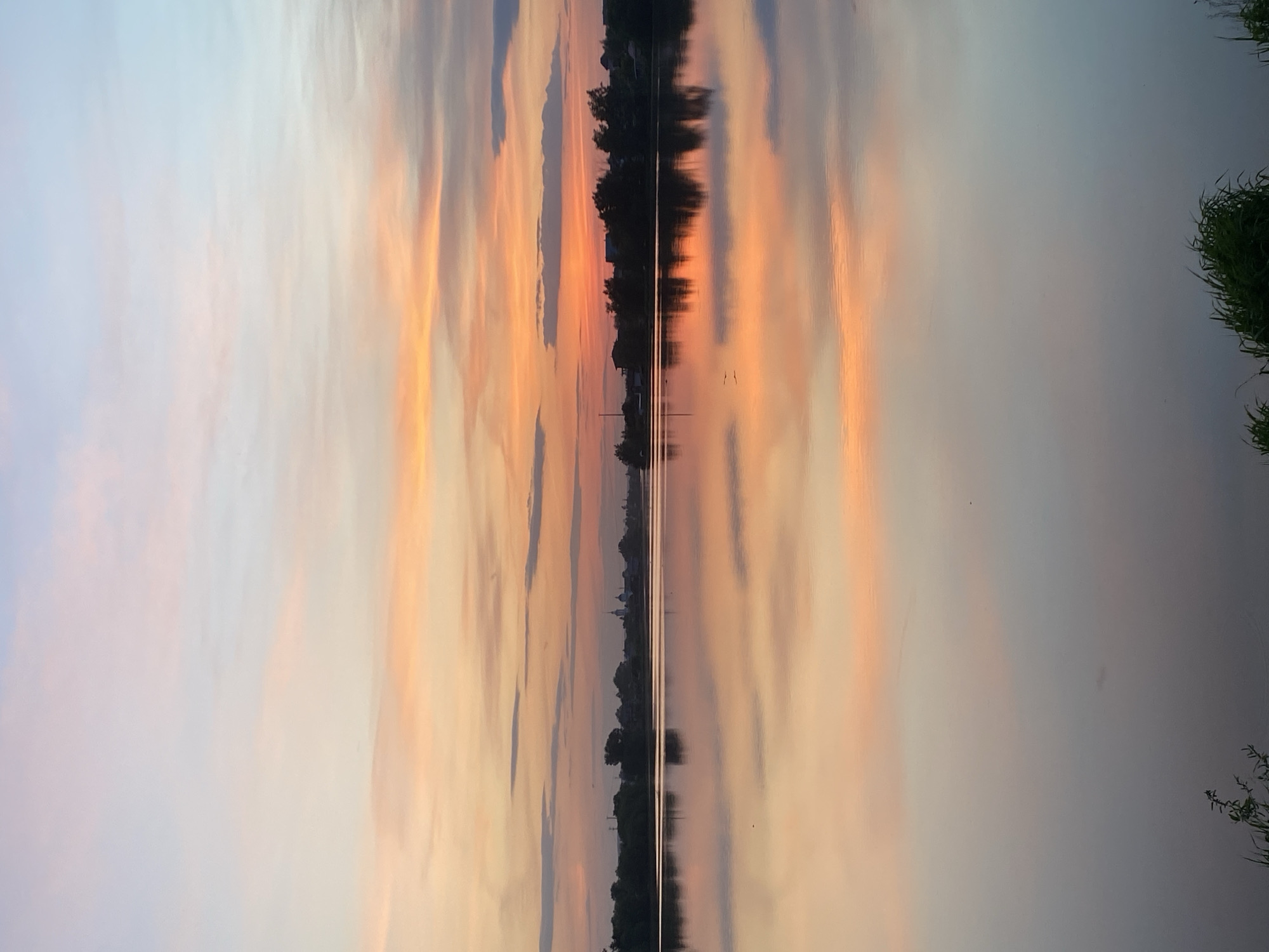 The image depicts a reflection of a sunset sky in a body of water. The sky features streaks of orange, pink, and blue hues. A thin line of trees, potentially on the shoreline, is visible in the middle of the image. Grass is present on the right side of the image, and there are some barely visible structures in the center of the image, which is the town Aramil, Russia.