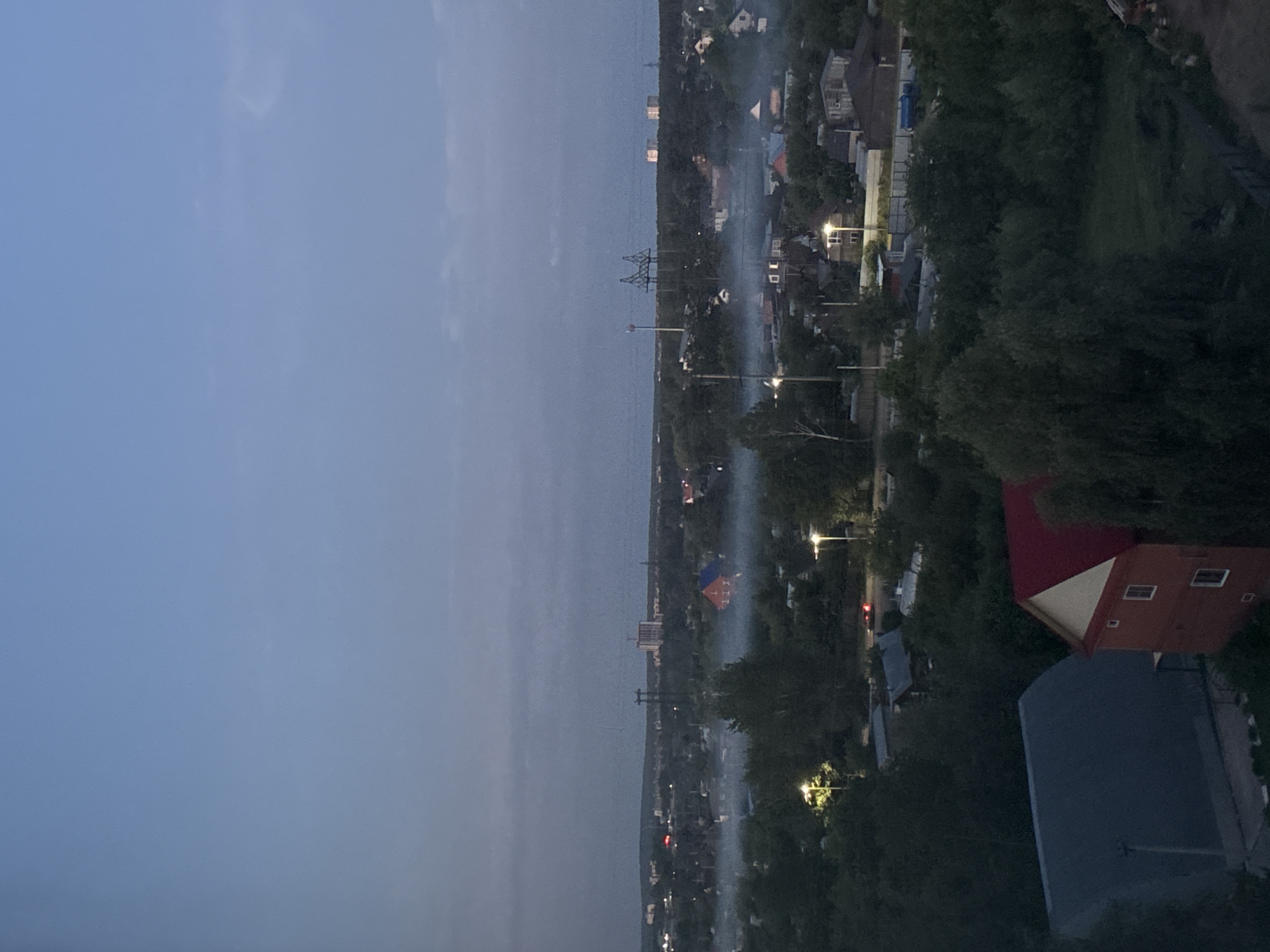 The image presents an aerial or high-vantage point view of a town or suburban area in Chaikovsky, Russia, captured during dusk or early evening. The scene is dominated by a hazy atmosphere, possibly due to mist, fog, or smoke.
The upper portion of the frame features a clear sky, transitioning from a light blue directly above the horizon to a deeper, greyish-blue towards the top, indicating the fading daylight. Below the horizon, the landscape is a blend of natural greenery and scattered urban development. Dense, dark green trees cover significant portions of the terrain, suggesting a lush environment.
Nestled among the trees are various buildings, primarily residential houses with distinct roofs, such as a prominent red-roofed house with white trim visible in the lower right. Streetlights punctuate the gathering darkness, casting bright yellow and white illumination onto roads and properties, highlighting areas otherwise obscured by shadow. A notable feature is the widespread, white, cloud-like mist or smoke that permeates the air, snaking horizontally across the middle and distant parts of the town, partially obscuring visibility and lending a mysterious quality to the scene.
Utility infrastructure is also visible, including several tall metal electricity transmission towers standing against the skyline in the middle distance. A red traffic light can be faintly observed in one section of the town. In the far background, the silhouette of more buildings stretches along the distant horizon, suggesting a wider settlement beyond the immediate view. No people or visible text are discernible. The overall atmosphere is calm and quiet, with the artificial lights beginning to dominate as natural light fades.