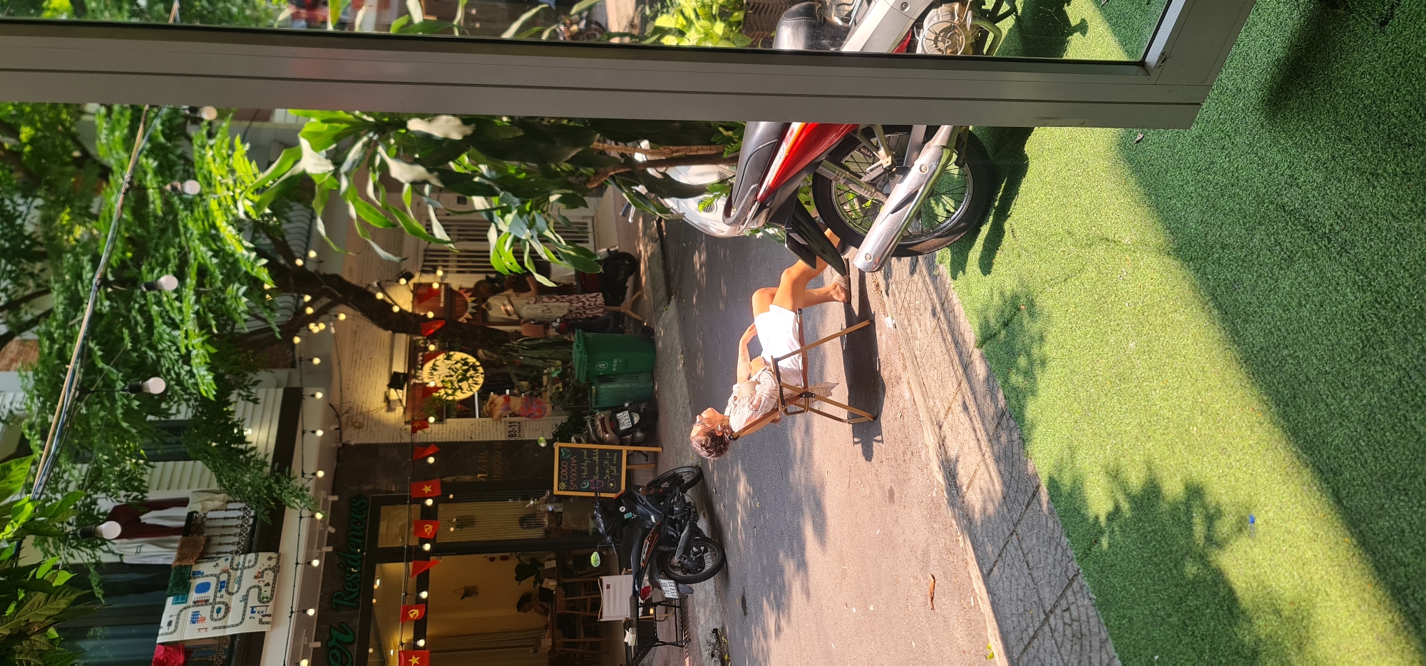 A medium shot of a young woman sitting on a folding chair on a city street. The woman is fair skinned with dark hair and is wearing a white top and white shorts. She is reclined in the chair and looking at something out of view. Behind her is a parked black scooter, green recycling bins, and a chalkboard sign. In the background is a storefront that reads "Residences" with decorative string lights. To her right is a parked red and silver motorcycle on a patch of artificial turf. The image is taken from the indoors looking through a window. The location is in Đà Nẵng, Vietnam, based on the red and yellow Vietnamese flags adorning the storefront.