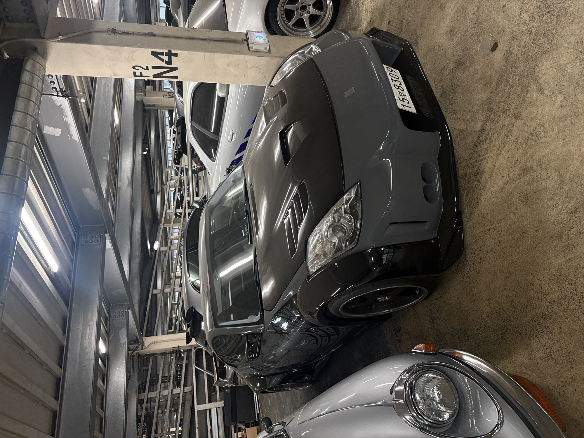 The image depicts an indoor parking structure, likely in the city of Seoul, South Korea. The structure is multilevel, constructed of gray steel beams and columns. A gray, customized sports car is parked near a column labeled "F2 N4". A white sedan is parked behind the sports car, and a silver classic car is partially visible in front. The lighting is provided by fluorescent lamps on the ceiling. A license plate on the gray car reads "15도 8309". The ground is concrete. No people are directly visible.