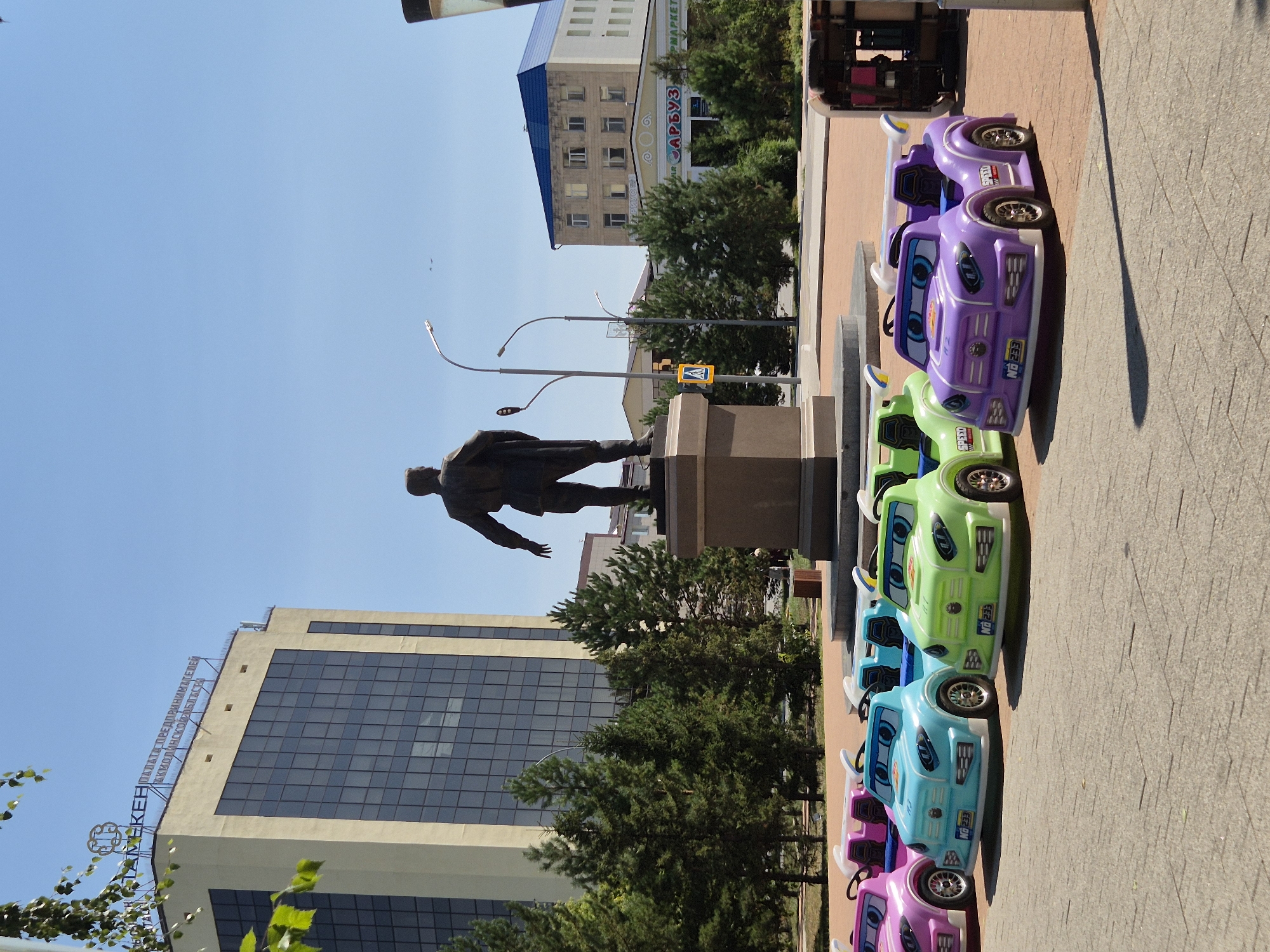 The image depicts an outdoor scene in the city of Kokshetau, Kazakhstan, under a clear blue sky. In the foreground, a row of brightly colored children's ride-on cars are parked on a paved area. The cars are purple, green, blue, and pink, and they are designed to resemble sports cars with cartoonish eyes on the front. One of the purple cars has a license plate that reads "NB 233".
Behind the cars, a large statue of a man in a suit, standing on a pedestal, is prominent. The statue faces away from the viewer, with its right arm extended. Further in the background, there are several buildings. To the left, a tall, modern building with a cream-colored facade and numerous dark windows is visible. A sign on top of this building reads "ПАЛАТА ПРЕДПРИНИМАТЕЛЕЙ АКМОЛИНСКОЙ ОБЛАСТИ" (Chamber of Entrepreneurs of Akmola Region). To the right of the statue, there is a smaller, lighter-colored building with a blue roof. In front of this building, a sign with Cyrillic lettering, including "САРБУЗ", is visible.
The area is also populated with lush green trees, suggesting a park or a public square setting. The overall impression is a sunny day in a city plaza, with a mix of modern architecture, historical elements like the statue, and recreational amenities for children.