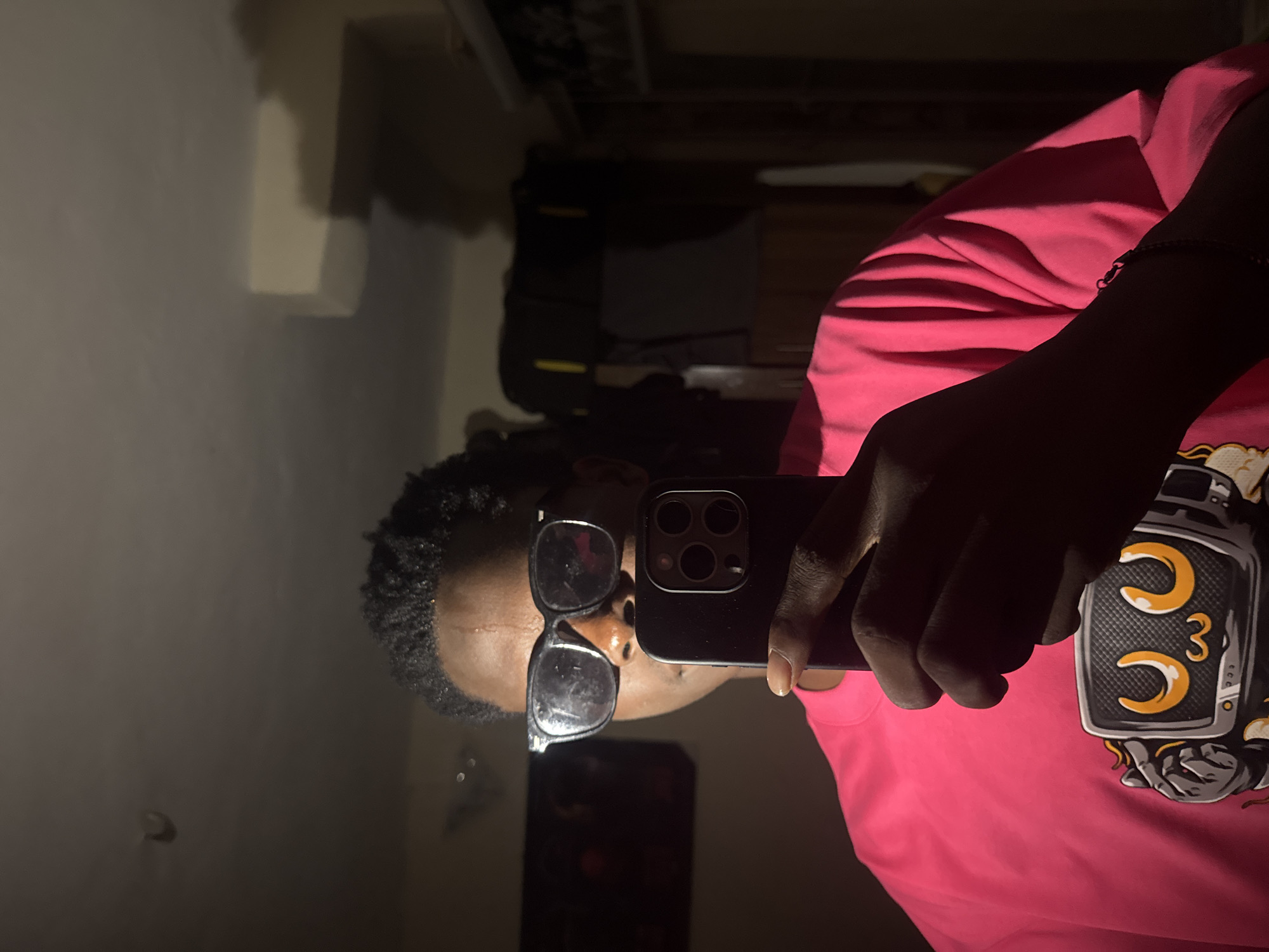 The image captures an individual taking a selfie indoors in Ihiagwa, Nigeria. The subject is a young person with dark, short curly hair, wearing black-framed glasses and a vibrant pink long-sleeve crew-neck shirt. The shirt features a graphic design of a robotic character with a screen for a face, glowing orange eyes, and mechanical hands. The individual holds a dark-colored smartphone, characterized by its prominent triple-lens camera module, vertically in their right hand, positioned to capture their reflection. A dark bracelet is visible on the subject's left wrist. The scene is illuminated by a strong, directional light source from the subject's left, creating stark highlights on the left side of their face, forehead, and the pink shirt, while casting the right side of the subject and the phone into deep shadow. The background is dimly lit and indistinct, revealing a dark, reflective surface (potentially a television or window) to the subject's left. To the subject's right, shadowy forms of furniture or shelving units are visible, along with a faint yellow detail on an object in the upper right. The ceiling is a light color, with a small, curved fixture discernible in the upper left quadrant. The environment appears to be a private room.