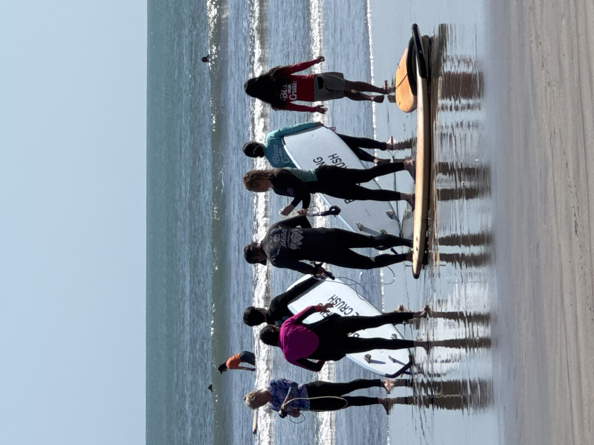 In the shallow waters of Kuta Beach, Bali, Indonesia, a group of people are participating in a surfing lesson. The water is calm with small waves rolling in, and the sky is a clear blue. Several individuals, dressed in wetsuits, stand on the wet sand and in the shallow water, holding surfboards. One person in a pink and black wetsuit is standing with a white surfboard. Another person in a black wetsuit is holding a white surfboard with text that appears to read "BLUE CRUSH". Further back, a person in an orange shirt is in the water, and two other individuals are visible in the distance on surfboards. The scene captures a typical day at this popular Indonesian surf spot.
