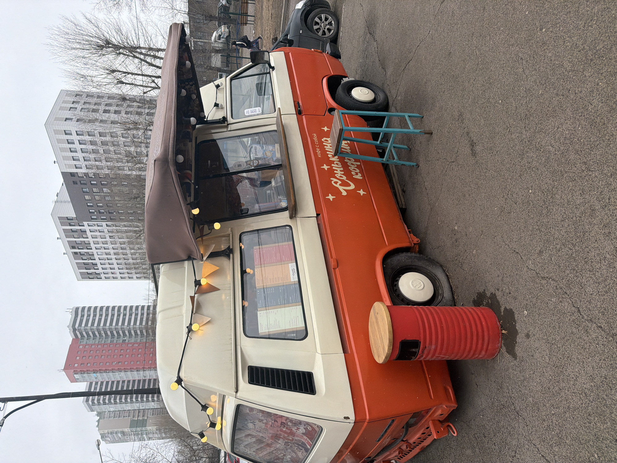 The image shows a stationary coffee truck in Krasnoyarsk, Russia. The vehicle is a two-tone orange and white vintage van, fitted with an awning and decorated with hanging lights and pennants. Cyrillic text reading "Солнышкина кофе с собой" is painted on the orange section of the truck. Next to the truck are a small blue step stool and a red metal barrel converted into a table. Several modern high-rise apartment buildings are in the background. The day appears overcast. The setting is likely a parking area or open space near residential buildings.
