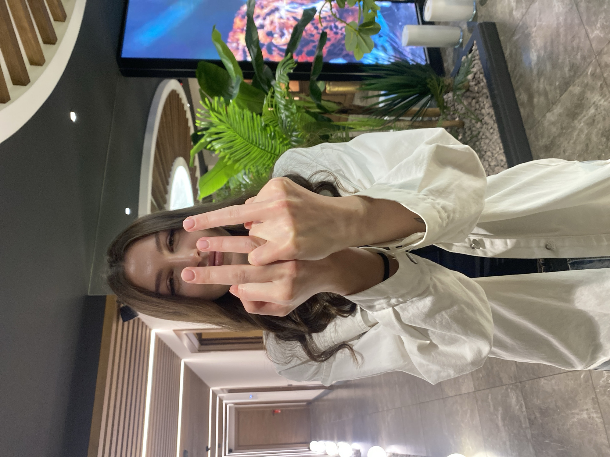 A young woman with long, wavy brown hair, wearing a white button-down shirt, stands in what appears to be a modern indoor space, possibly a hotel lobby or a commercial building. She is looking directly at the camera with a neutral expression and is holding both of her hands up, displaying the middle finger on each hand. The background shows a long hallway with illuminated panels on the ceiling and walls, and to her right, there is a large potted plant and a digital screen displaying an image. The floor is tiled. The context of Votkinsk, Russia, is not visually discernible from the image.
