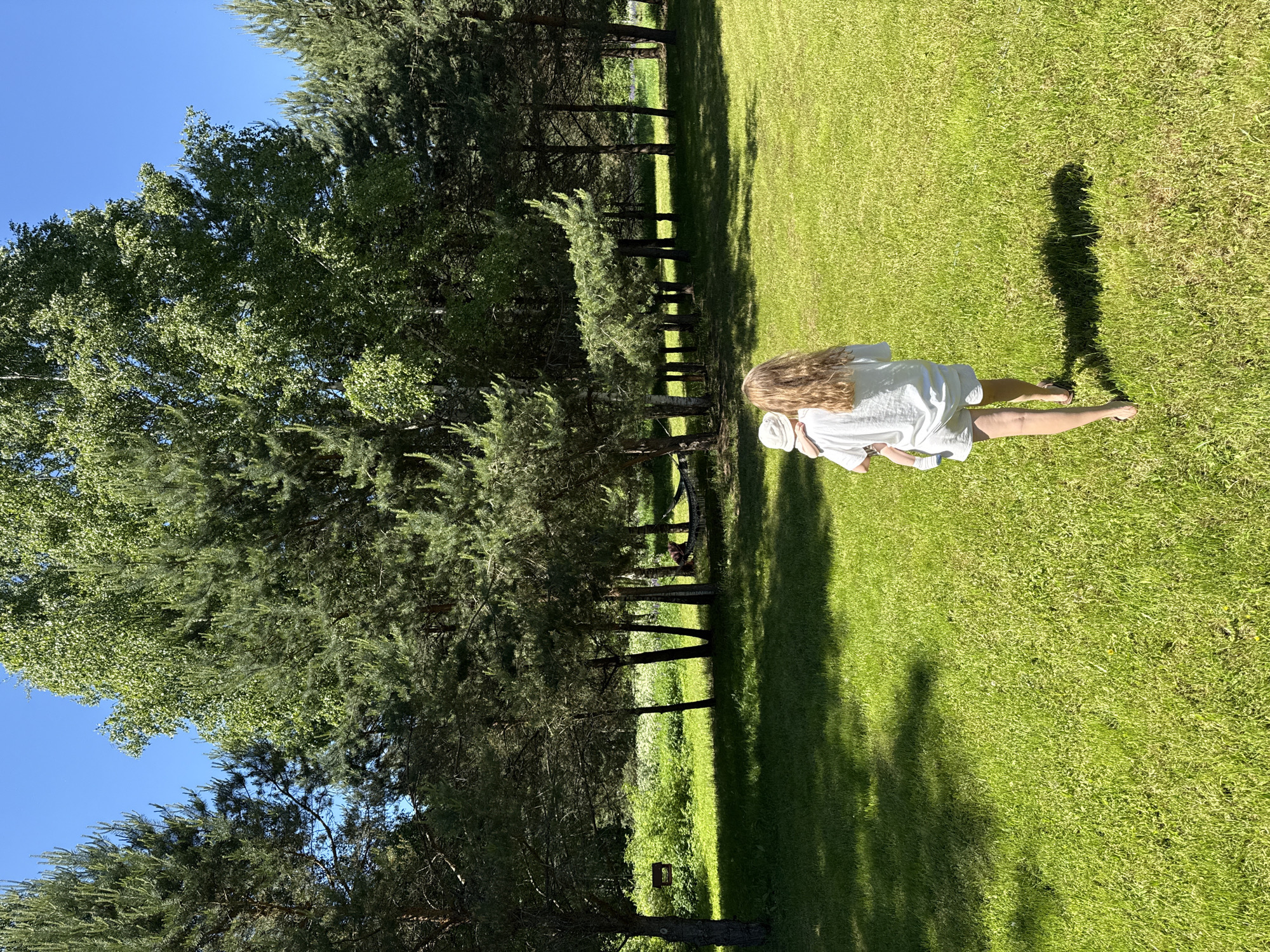 In Копнино, Russia, a bright, sunny outdoor scene displays a wide expanse of vibrant green grass, partially shaded by trees. An adult, viewed from behind with long blonde hair, wears a white t-shirt, white shorts, and dark sandals, carrying a baby. The baby, also seen from behind, wears a white bucket hat and light-colored clothing. Both are walking away from the camera, across the lawn, towards a dense treeline in the background. Strong sunlight creates distinct shadows on the grass. The treeline comprises numerous tall evergreen pine trees interspersed with deciduous trees, including a prominent birch tree with light bark. Within the treeline, a dark rectangular wooden sign is visible hanging from a tree, and a person is relaxing in a dark-colored hammock strung between two trees. A clear, bright blue sky is visible above the trees.