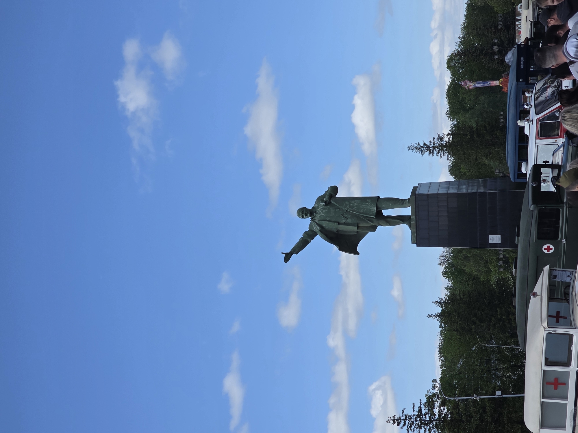 A statue of Lenin stands in front of a blue sky with scattered clouds. The statue is dark in color and depicts Lenin in a coat, with one arm outstretched. Behind the statue, there is greenery and vehicles, including what appears to be an ambulance, and some people can be seen. The location is in Ufa, Russia.
