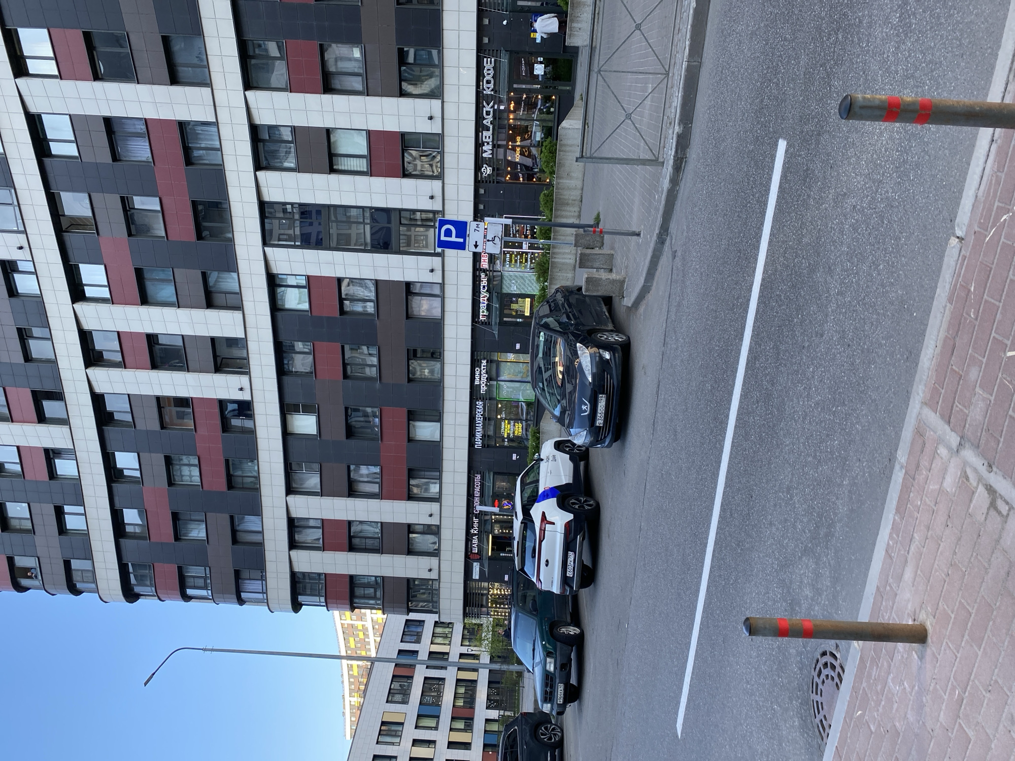 The image shows a street scene in Murino, Russia. A modern, multi-story building dominates the left side, featuring a pattern of dark gray, white, and maroon panels with windows. At street level, businesses like "M:BLACK KOFE" and "ШАВА КИНГ САЛОН КРАСОТЫ" are visible. A "P" parking sign and a sign advertising "Вино продукты" (wine products) are also present. Several cars are parked along the street, with license plates indicating they are registered in the Saint Petersburg region of Russia, such as "E36600 198", "C603PK 797", and "P964KA 50". A white lane marker separates the parking area from the street. Metal bollards with red stripes stand on the right, marking the edge of a brick-paved area. The sky is clear blue.