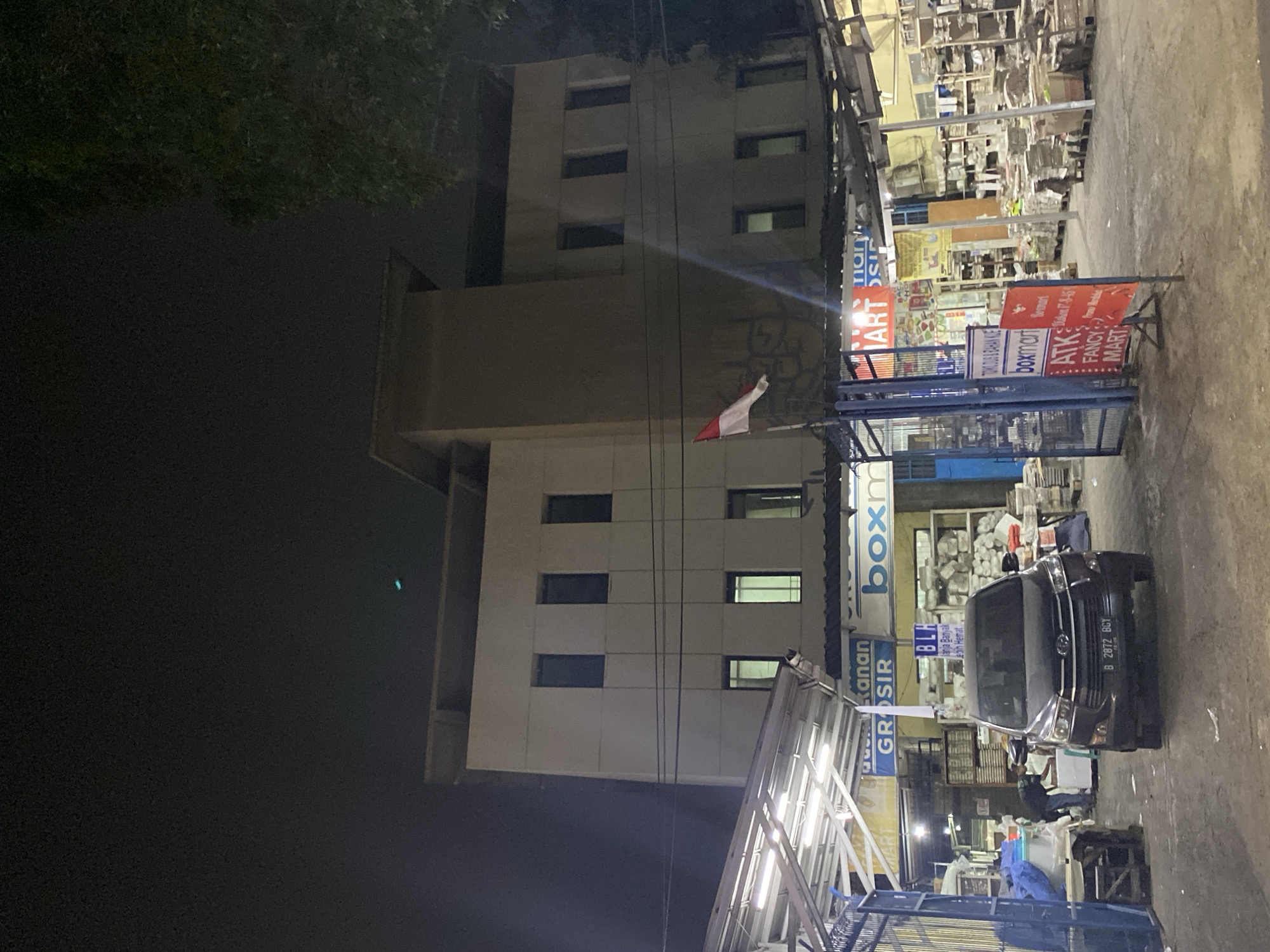 The image shows a commercial building at night, likely a warehouse or a multi-store complex, located in Tangerang, Indonesia. A dark-colored SUV is parked in front of the building. The building has several floors with numerous windows. Signs visible on the building and in front of it indicate businesses such as "GROSIR" (Wholesale), "BOXMAN," and "ATK FANCY MART." The area appears to be a retail or wholesale district. A small Indonesian flag is visible on a pole. The lighting suggests artificial light from within the shops and streetlights. Trees are visible on the right side of the image.