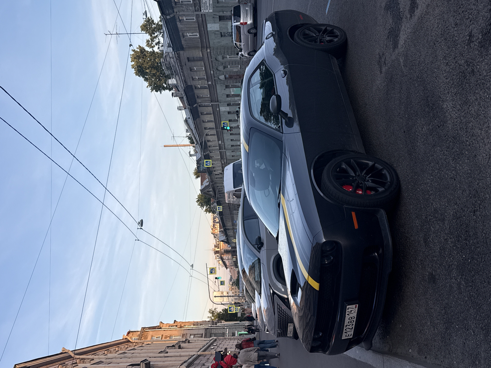 A dark-colored Dodge Challenger with yellow racing stripes on the hood and roof is parked on the right side of a cobblestone street in Saint Petersburg, Russia. The car's license plate is visible and reads "E123BB 147 RUS". In the background, a row of historic buildings with ornate facades lines the street. Pedestrians are walking on the sidewalk to the left, and other vehicles, including a light-colored SUV and a white van, are also present. Traffic lights and pedestrian crossing signs are visible, indicating an intersection. The sky is clear with a few wispy clouds, and the lighting suggests it is late afternoon or early evening.
