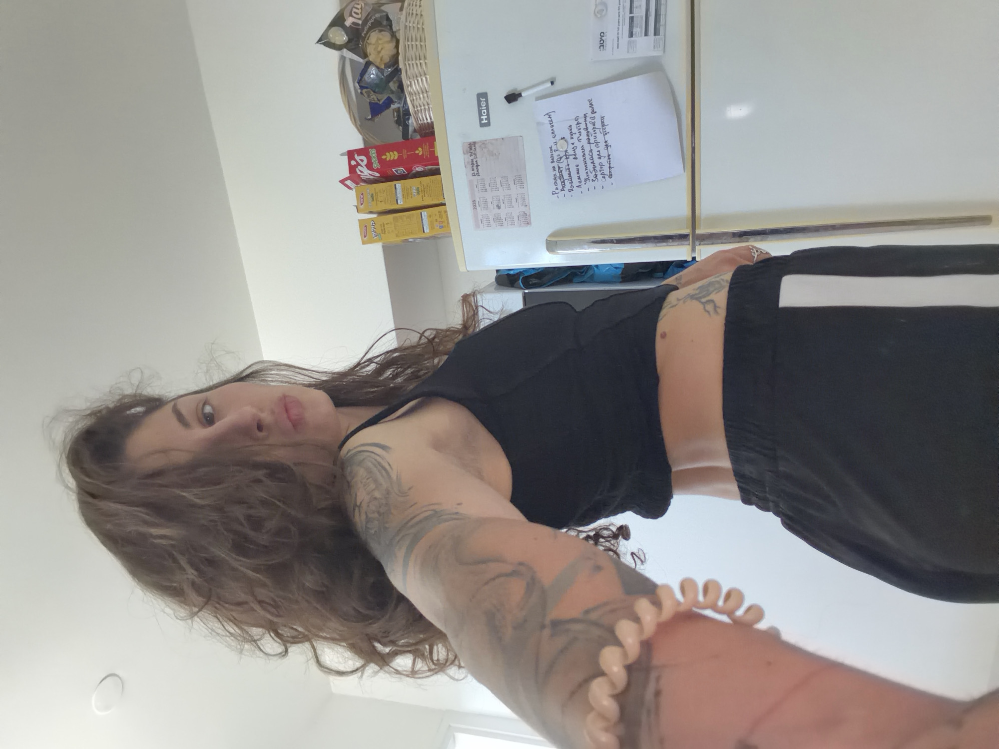 A young woman with wavy brown hair and arm tattoos is taking a selfie in a kitchen in Kiryat Bialik, Israel. She is wearing a black crop top and black pants with a white stripe. The background includes a white refrigerator with a shopping list attached, a basket of snacks, and boxes of cereal. The list is written in Hebrew and contains shopping items and to-do tasks.