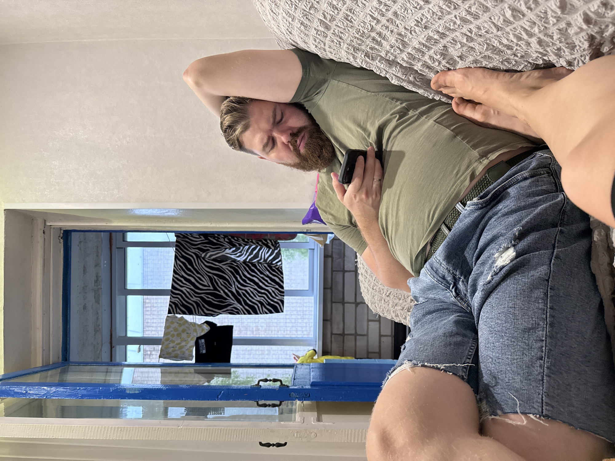 A man is lying on a textured gray couch, holding a black smartphone in his right hand. He is wearing a faded olive green t-shirt and ripped denim shorts. His eyes are closed, and his head is tilted back, resting on his right arm which is bent behind his head. A thick brown beard covers his chin and jawline. The couch is positioned in an indoor setting, with a partially open blue door visible to the left of the frame. Beyond the door, laundry is hanging on a line, with various items including a yellow patterned garment and a black and white striped fabric. The background appears to be a balcony or porch area with windows and brickwork. The flooring visible through the doorway is tiled. The overall lighting suggests an indoor environment, possibly during the daytime given the light coming through the windows.