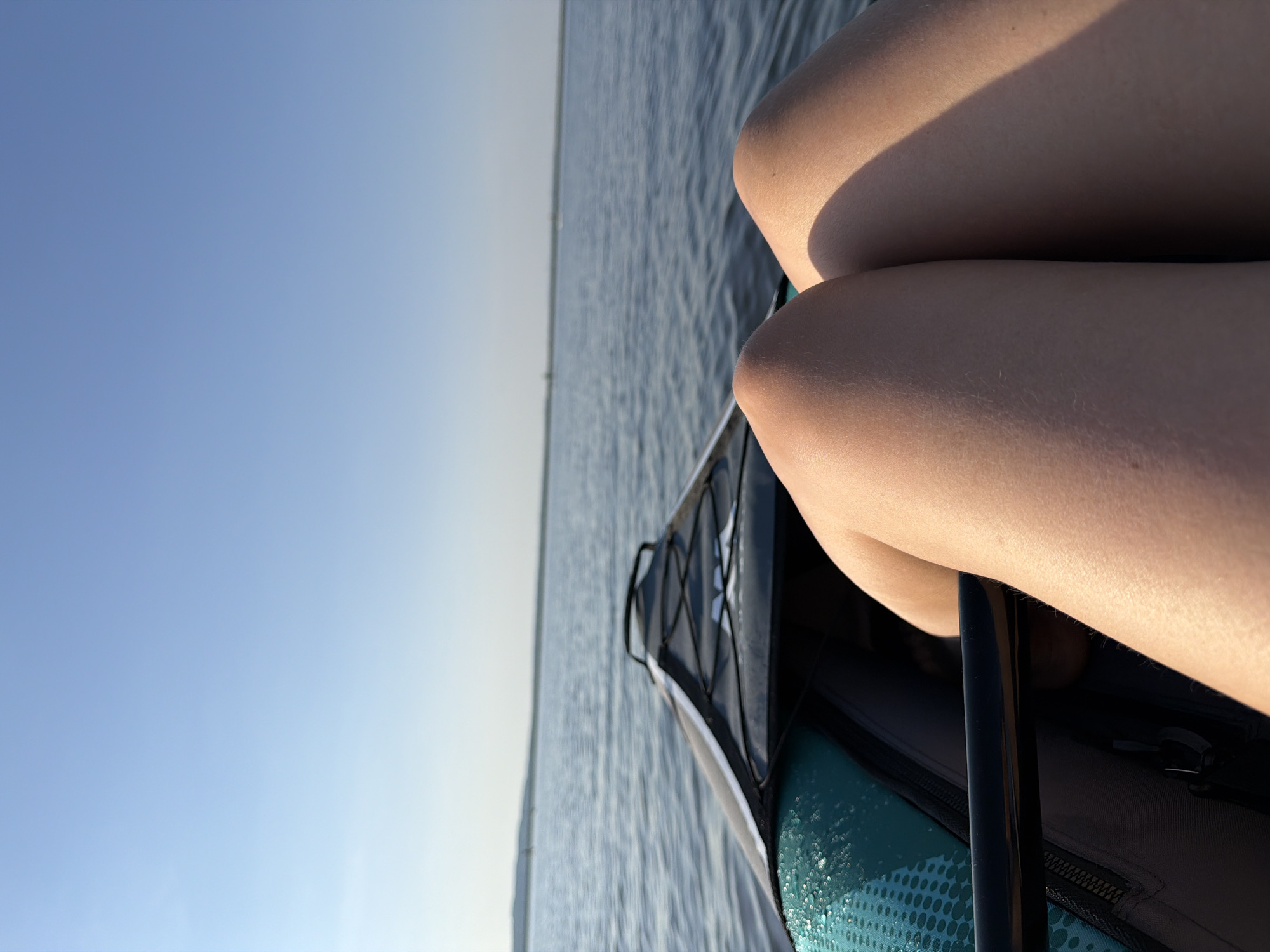 The image depicts a view from a kayak on a body of water. The sky is clear and blue. The water surface shows small ripples. The knees of a person are visible in the right side of the frame, suggesting they are sitting in the kayak. A portion of the kayak is also visible, including its storage area, bungee cords, and frame. I cannot verify the location.
