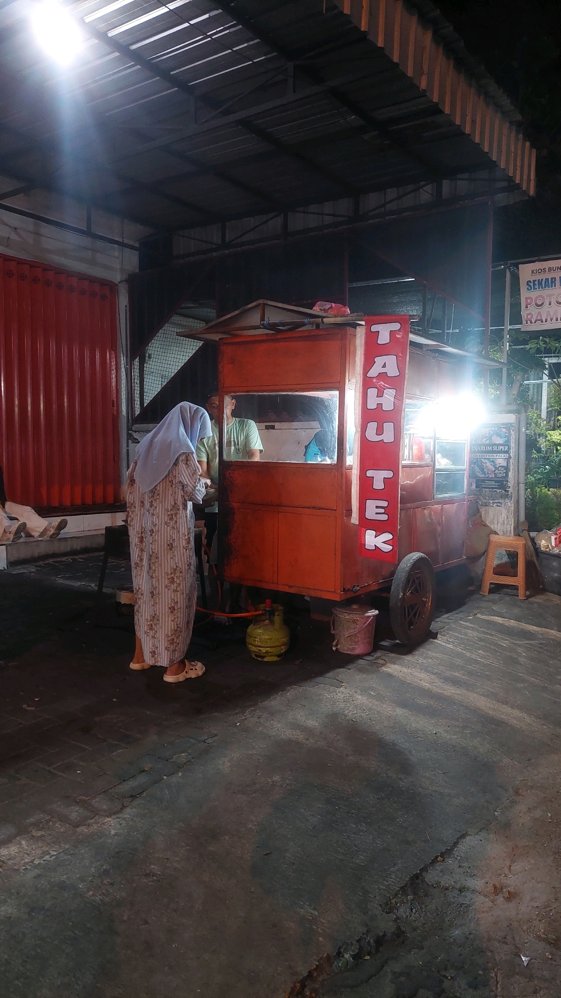 The image depicts an outdoor scene at night, likely in Banyudono, Indonesia, featuring a food cart. The cart, painted orange, has a vertical red banner with white lettering that reads "TAHUTEK". A person wearing a light blue hijab and a patterned dress is standing in front of the cart, appearing to be interacting with someone inside. Another person, wearing a light green t-shirt, is visible behind the counter of the cart. A green gas cylinder is placed on the ground next to the cart, and a pink bucket is also visible. The ground is paved with textured concrete, and there are some shadows cast by artificial lighting. In the background, there is a red corrugated metal structure and a roof with visible beams and lighting. A small orange stool and some foliage are also present. A sign with text is attached to the wall in the background.