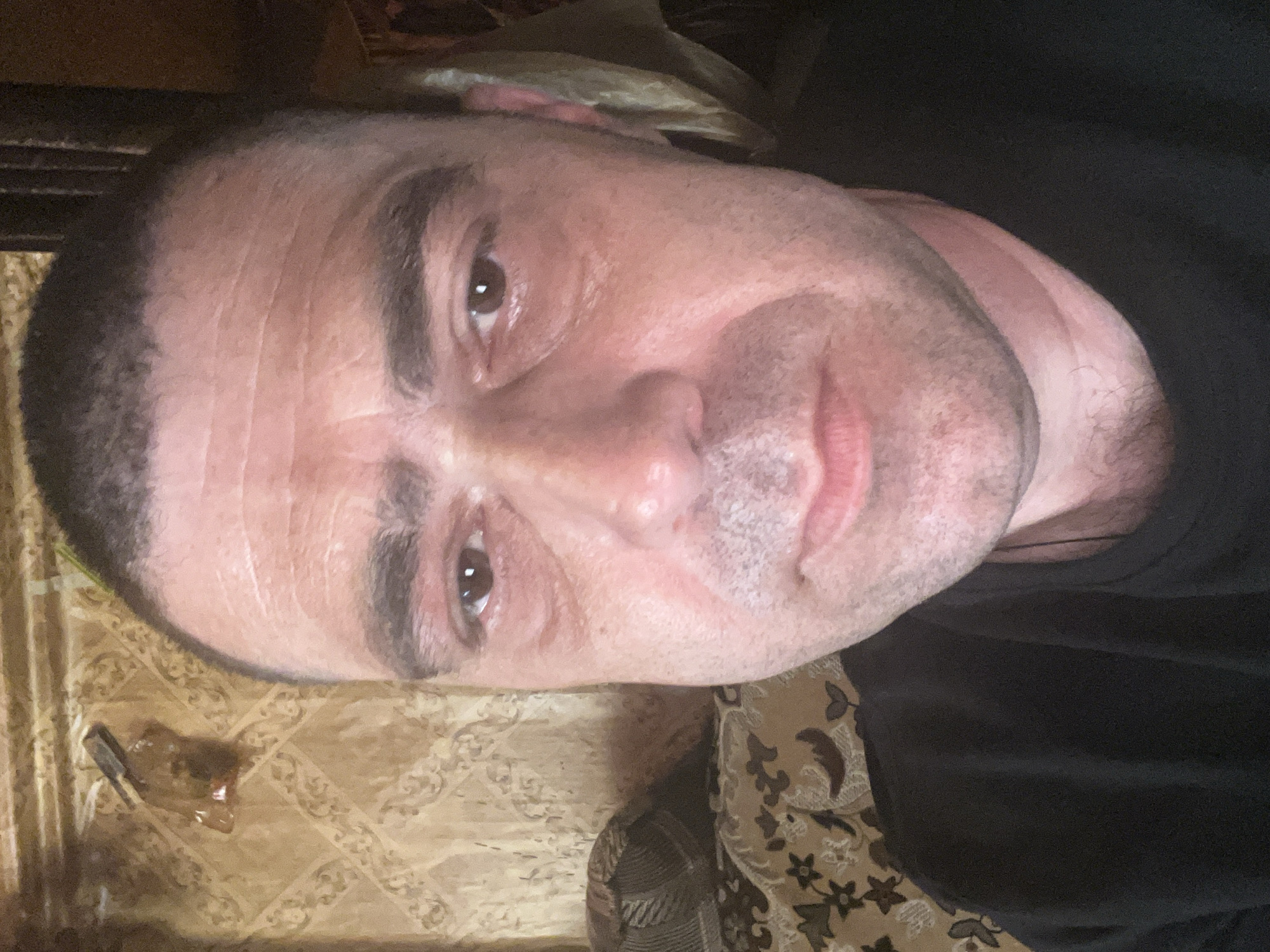 The image is a close-up, rotated selfie of a man. He is looking towards the camera with a slight smile. The man has short, dark hair, and his face shows some wrinkles and stubble. He is wearing a dark t-shirt. The background appears to be indoors, with a patterned carpet or rug visible on the floor. To the left of the man, there is a small object that looks like a plastic bag containing something brown. The lighting suggests it might be daytime, but there are no strong indicators of the time of day or weather. The overall impression is a casual, unposed photograph.
