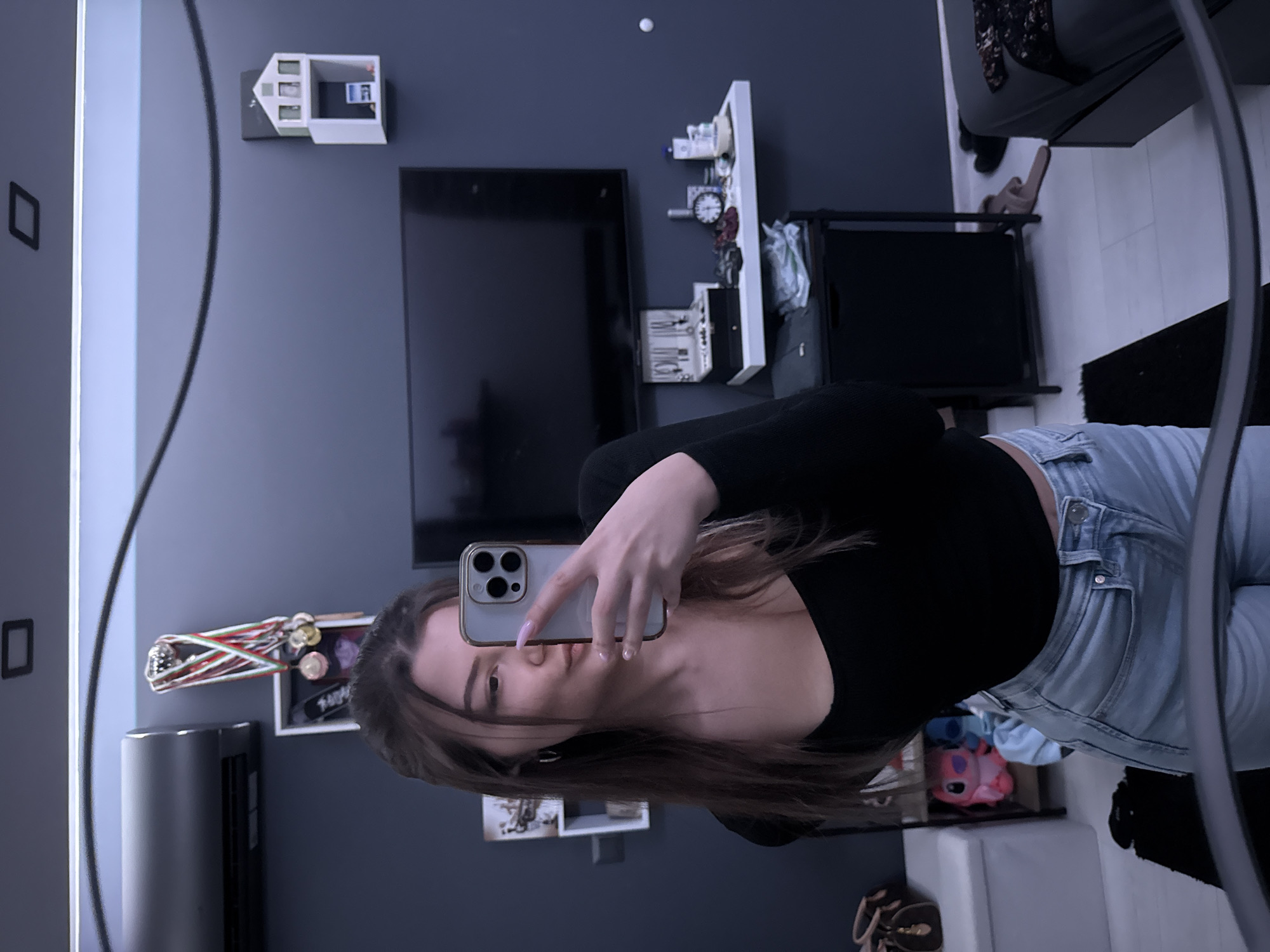 A mirror selfie, showcasing a young woman in a black long-sleeve shirt and light-wash jeans, holding her smartphone to capture the reflection. Her hair is brown, and she is positioned in front of a wall-mounted TV and various shelves containing decorations and personal items. The room has a modern aesthetic with a dark gray wall color. The location is likely a residential interior, potentially in Sofia, Bulgaria, though the specific city is difficult to ascertain with certainty from visual evidence alone.