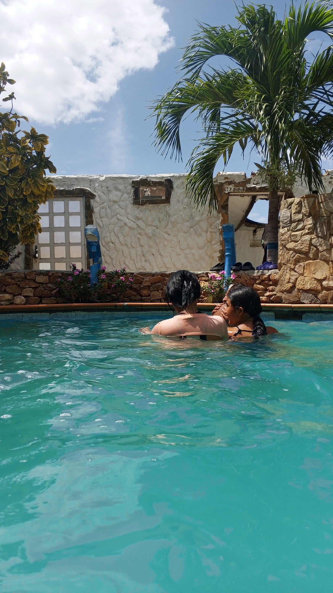 Two individuals are immersed in the turquoise water of a pool. The person on the left has their back to the viewer, with dark hair. The person on the right is facing the other individual. Behind the pool is a low stone and white wall. A window with square panes is on the left, with vegetation on the side. A partial palm tree is visible on the right. The location is Unknown, Unknown.