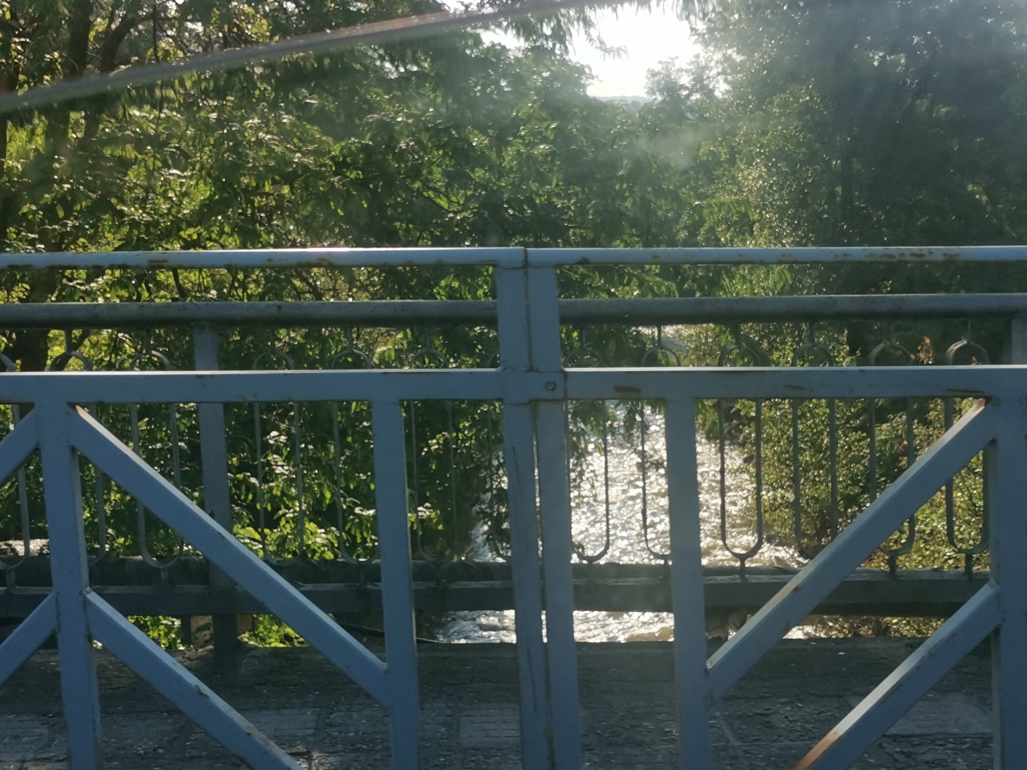 The image presents an outdoor scene viewed from behind a metal fence, likely from a bridge or elevated platform, overlooking a body of water and dense foliage.
In the foreground, a sturdy light blue-grey metal fence or railing dominates the lower half and extends horizontally across the entire frame. It consists of multiple horizontal bars and a network of vertical and diagonal supports, forming geometric sections. Decorative, looped or curved metal elements are visible between the lower horizontal bar and the ground surface. Some signs of wear and minor rust are apparent on the fence.
Through the openings of the fence, a sparkling body of water, likely a river or stream, can be seen below. Its surface is highly reflective, with numerous bright patches indicating direct sunlight hitting the water, creating a dazzling, rippling effect.
Beyond the water, the background is filled with lush, vibrant green trees and dense vegetation. Sunlight filters through the leaves, creating bright highlights and dappled light patterns within the foliage, suggesting a sunny day. The overall impression is one of natural abundance and tranquility.
The time of day appears to be sunny daylight, possibly morning or late afternoon, given the intense reflections on the water and the way light penetrates the tree canopy. There are no people visible, nor any discernible activity taking place. No text is visible in the image. The setting evokes a serene natural environment, fitting the provided context of Kostenets, Bulgaria.