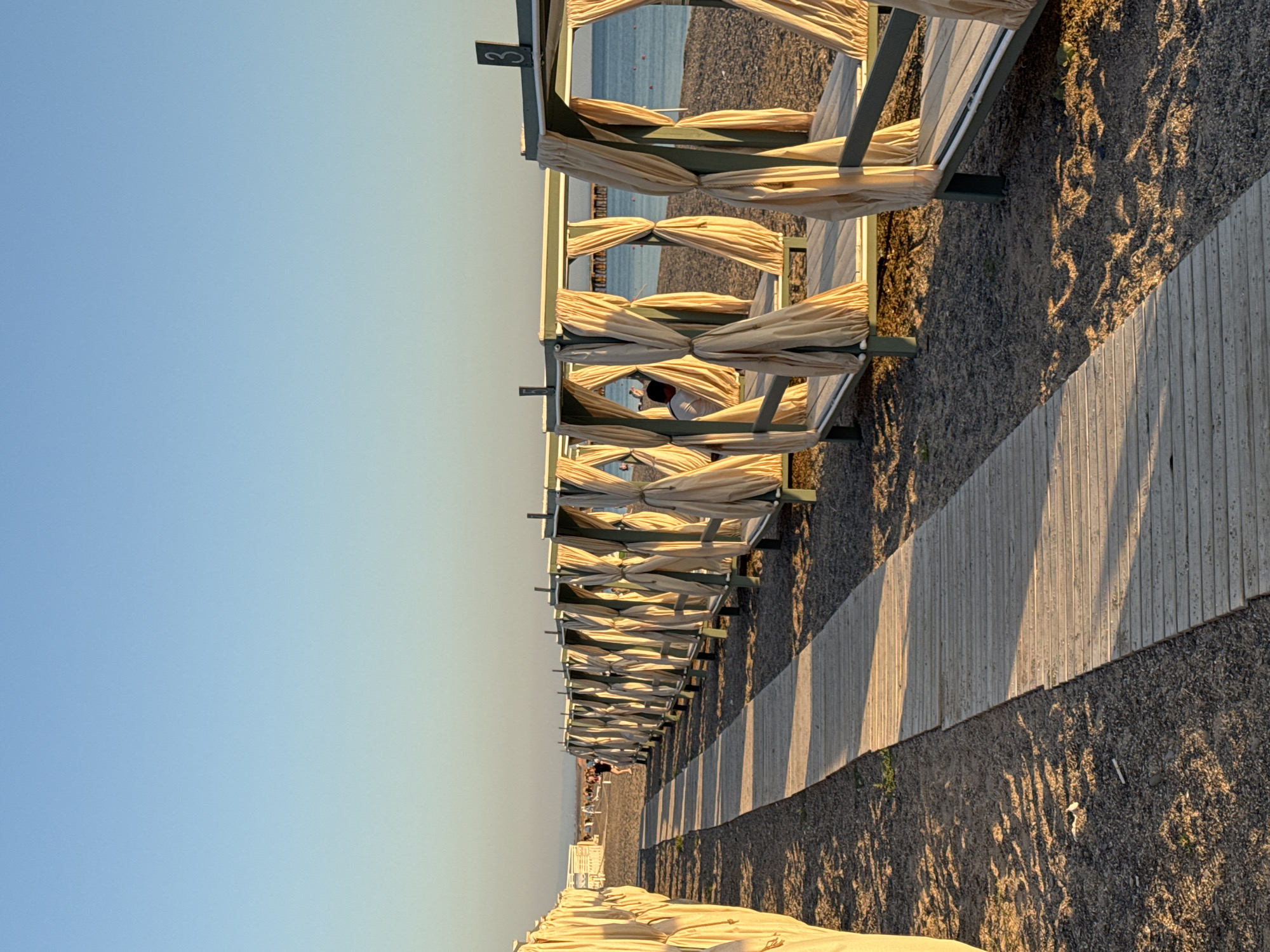 A low-angle, eye-level shot captures a wooden boardwalk leading down a sandy and pebbly beach toward the sea. The boardwalk is positioned to the left of the frame, with its wooden planks casting long shadows in the late afternoon sun. To the right of the boardwalk, a row of cabanas with light-colored curtains is visible. The cabanas are arranged linearly, extending into the distance. Some of the cabanas have the number "3" visible on them. In the background, the calm sea meets the horizon under a clear, light blue sky that transitions to a lighter shade near the horizon. Several people are visible on the beach in the distance, some near the cabanas and others further down the shore. One person is seated inside one of the cabanas. A pier or structure extends into the sea from the distant shoreline. The overall scene suggests a peaceful beach setting during golden hour.