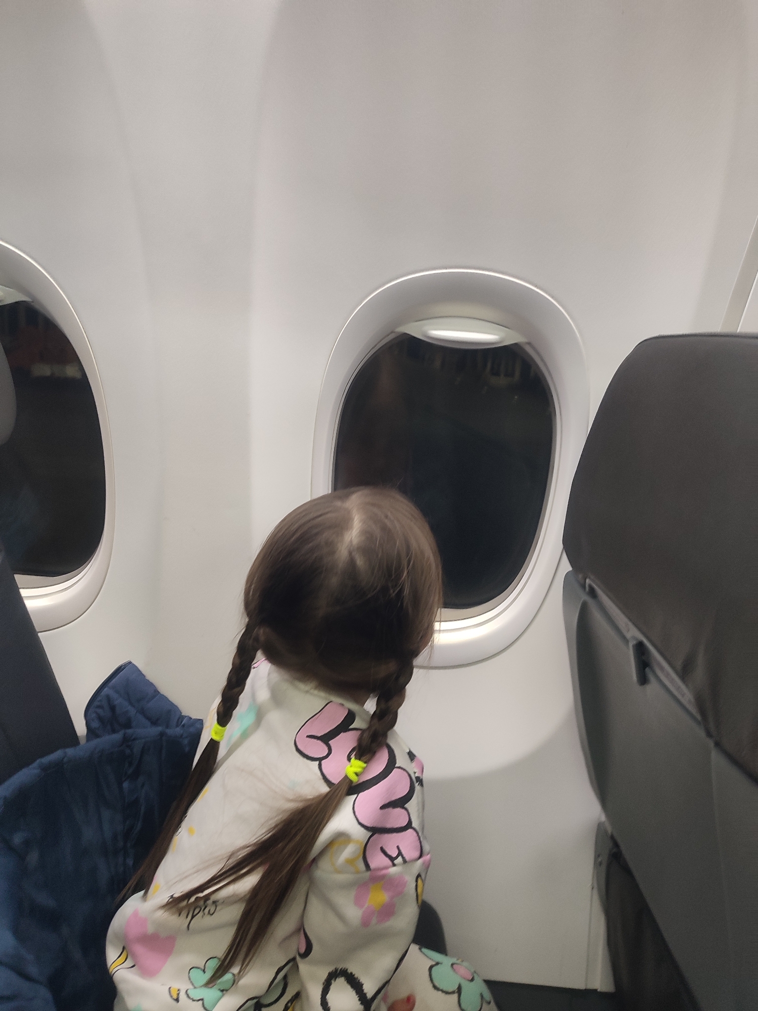 A young child is observed from a rear-view perspective inside an airplane cabin, likely located at an airport in Izhevsk, Russia. The child, with long, dark brown hair styled into two braids secured by neon yellow elastic bands, is wearing a light-colored garment patterned with various pastel shapes including hearts and flowers. The child is leaning forward, looking out of a circular aircraft window. The view through the window is dark and largely indistinct, showing only faint, blurry lights suggestive of an external ground environment at night. To the child's left, a dark blue quilted jacket or blanket is visible on an adjacent seat. The upper portion of a dark grey aircraft seat back is partially visible on the right side of the frame. The interior walls of the cabin are white or light grey.