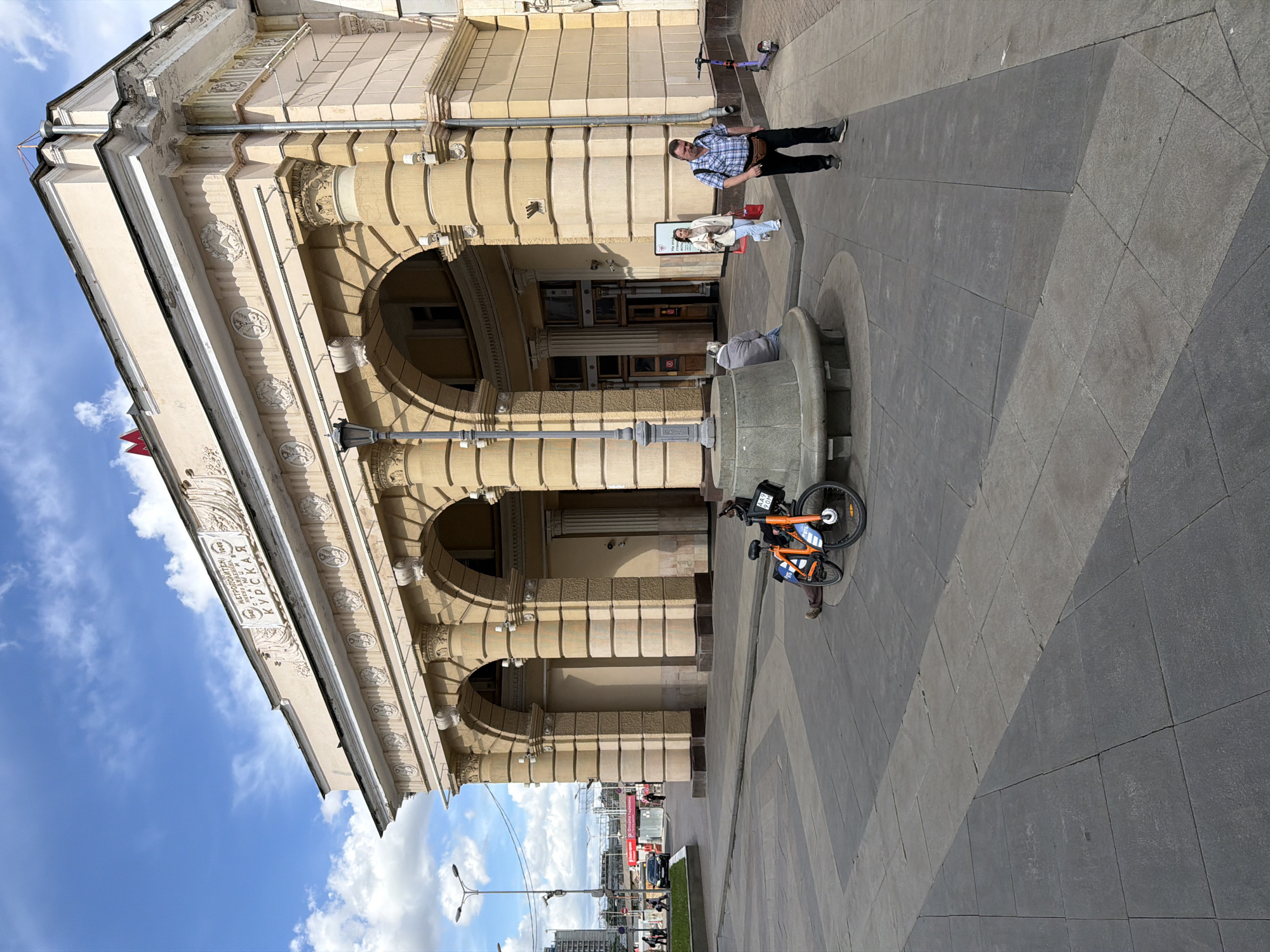 Here is a description of the image:
The photo captures a street scene in front of a grand, ornate building, possibly a metro station judging by the signage. The building has a facade with arched windows and columns, suggesting classical architecture. A bicycle is parked near a stone structure that could be part of a fountain or public art installation. Several people are visible: a man standing near the entrance, someone sitting on the edge of the stone structure, and others in the background, including a couple standing near a sign. The pavement is made of large, square tiles. The sky is visible with a few clouds. Construction work is visible in the background.