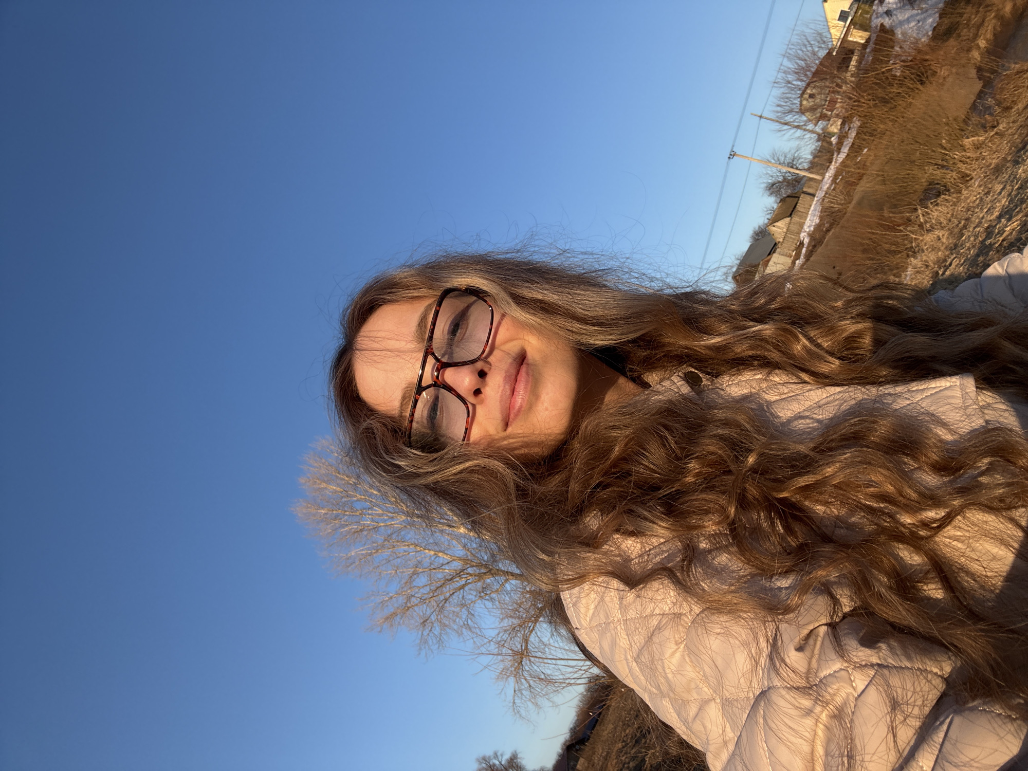 A photograph shows a young woman with long, wavy, light brown hair and glasses. She is wearing a light-colored, quilted jacket. The background includes a clear blue sky and a landscape with dry vegetation, trees, and buildings in the distance, suggestive of an outdoor location in Chaikovsky, Russia. The photograph appears to have been taken in the daytime, with the sun low in the sky casting long shadows. The image is taken from a slightly angled, low perspective.