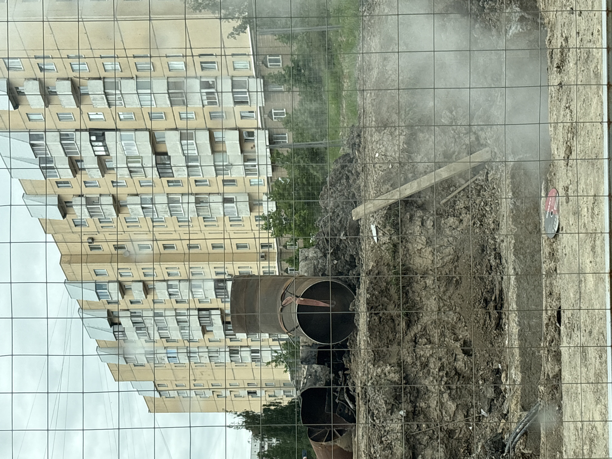 The image shows a construction site in Saint Petersburg, Russia. A large metal pipe lies in the dirt of an excavated trench. The building in the background is reflected in a window or some other reflective surface. A wire fence separates the viewer from the site. Visible in the background are residential buildings and trees. A grinding wheel sits on the ground to the right of the hole. A piece of wood lies atop a pile of dirt within the trench. The overall scene suggests an active construction project involving underground utilities.