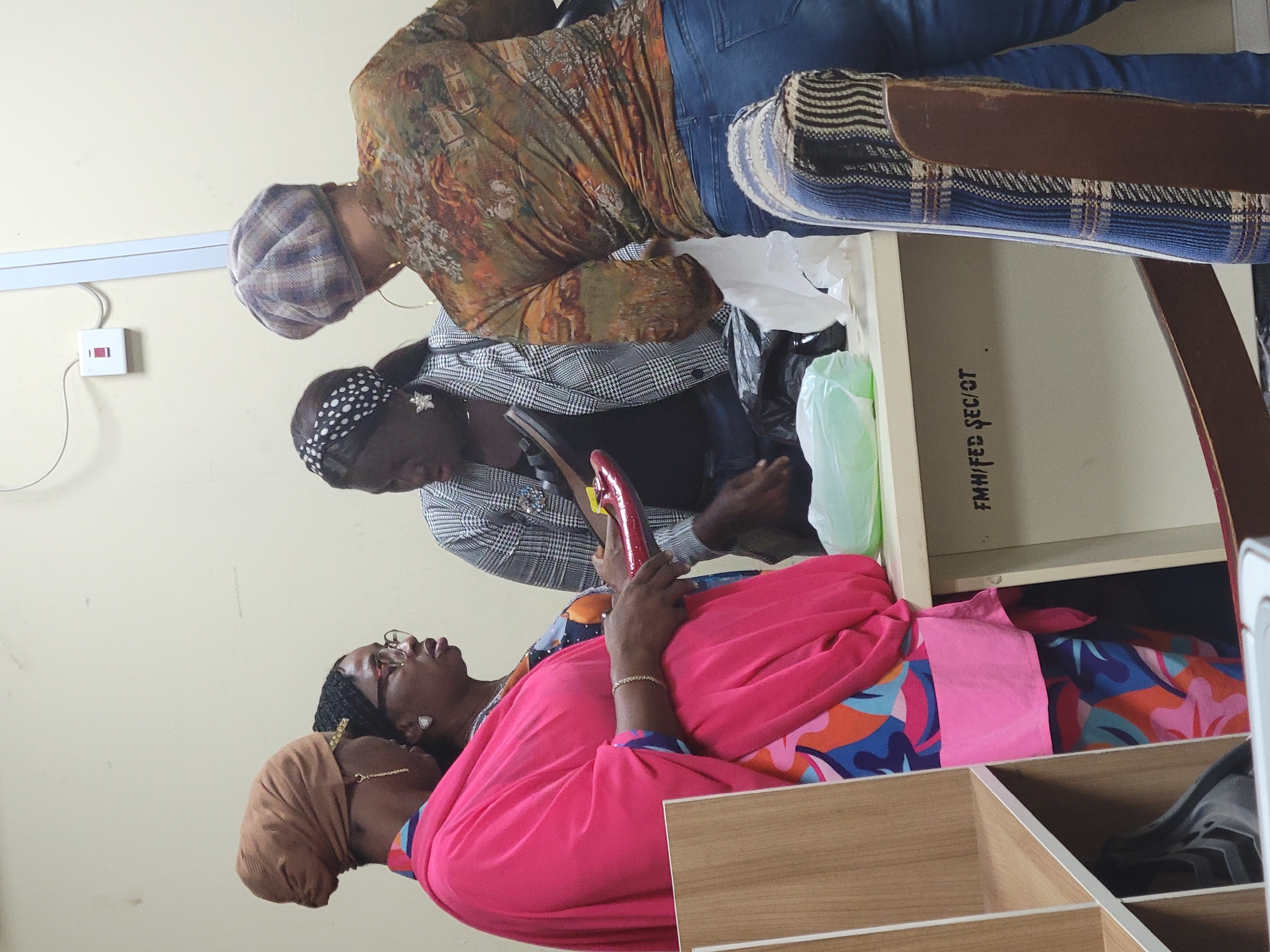 Three women are gathered around a shelf unit. One woman, wearing a brown head covering and a bright pink top, is in the foreground. Above her, another woman wears glasses and a black head covering. The third woman is at the top of the frame wearing a patterned shirt, jeans, and a grey cap. The shelf unit contains items wrapped in plastic. The location is Abuja, Nigeria.